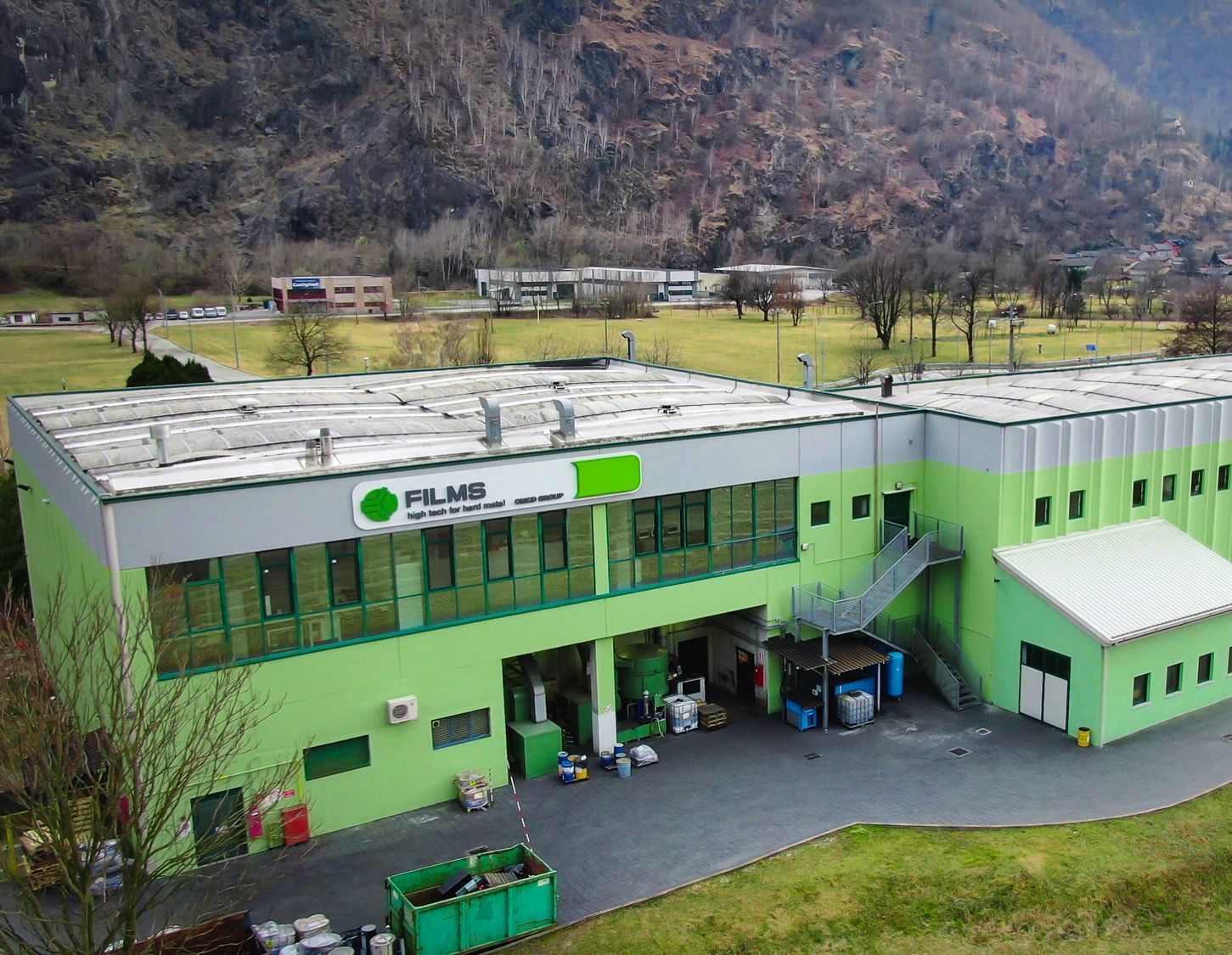 Veduta aerea di un edificio industriale di colore verde brillante e grigio, incastonato ai piedi di una montagna boscosa.