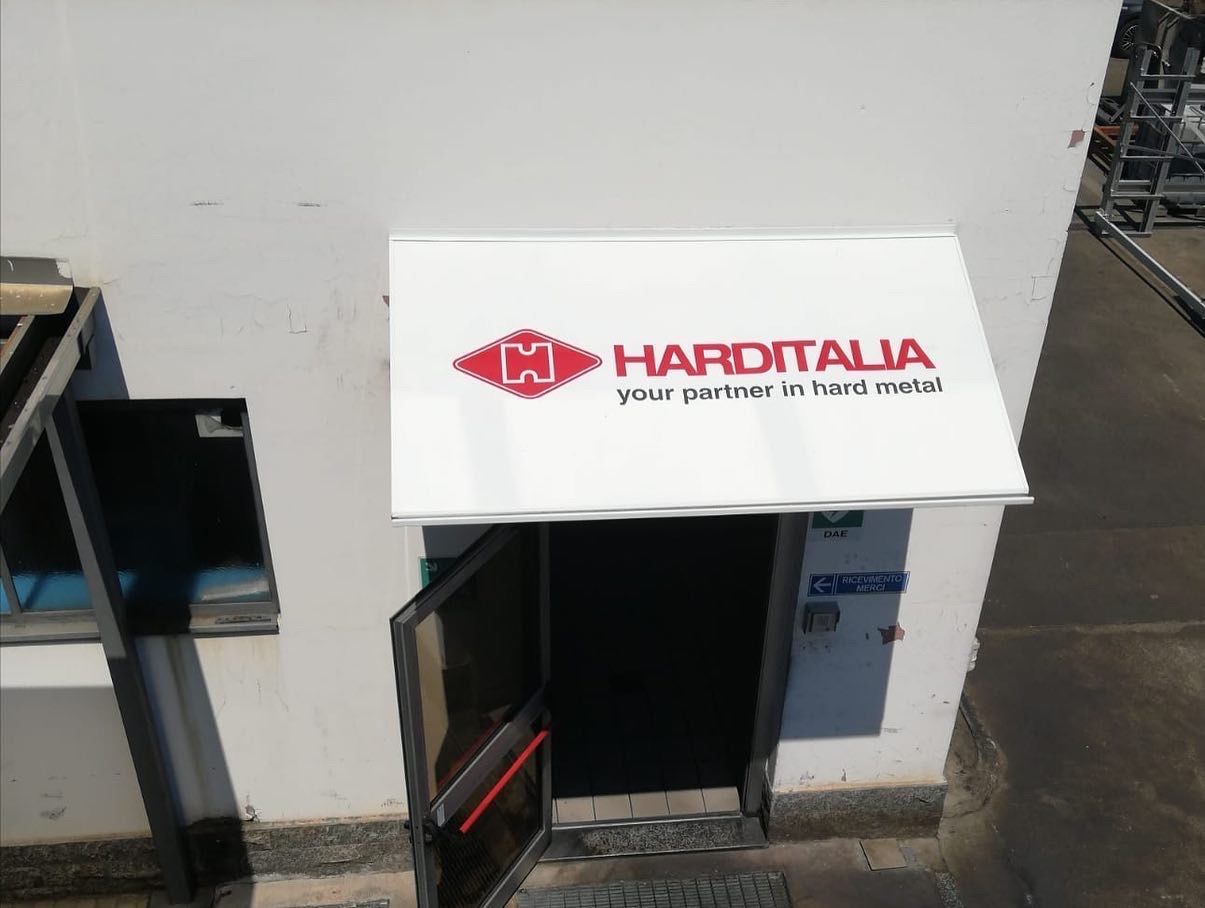 Un portone aperto con una tenda da sole bianca su cui sono stampati il ​​logo e lo slogan rossi di Harditalia, montata su una parete esterna bianca.