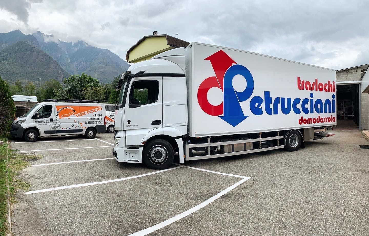 Un grande camion bianco per traslochi e un furgone bianco più piccolo parcheggiati su un piazzale asfaltato con le montagne sullo sfondo.