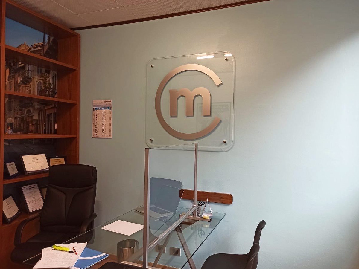 Un ufficio professionale con scrivania in vetro, sedie ergonomiche, libreria in legno e insegna con logo su una parete azzurra.