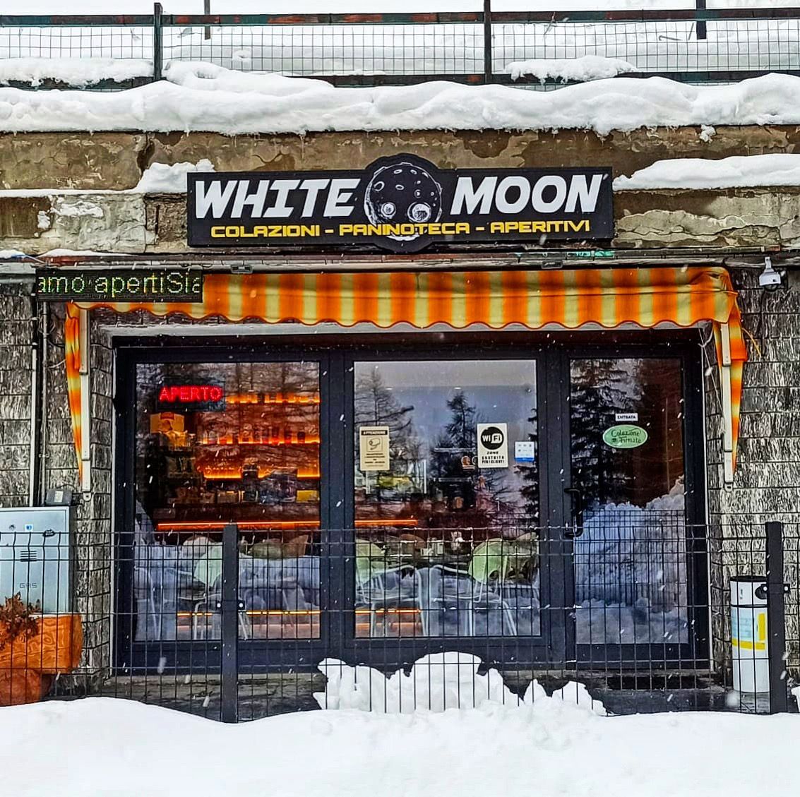 La facciata del caffè White Moon, con tenda da sole a strisce arancioni, insegna e porte a vetri, si staglia su un paesaggio innevato.