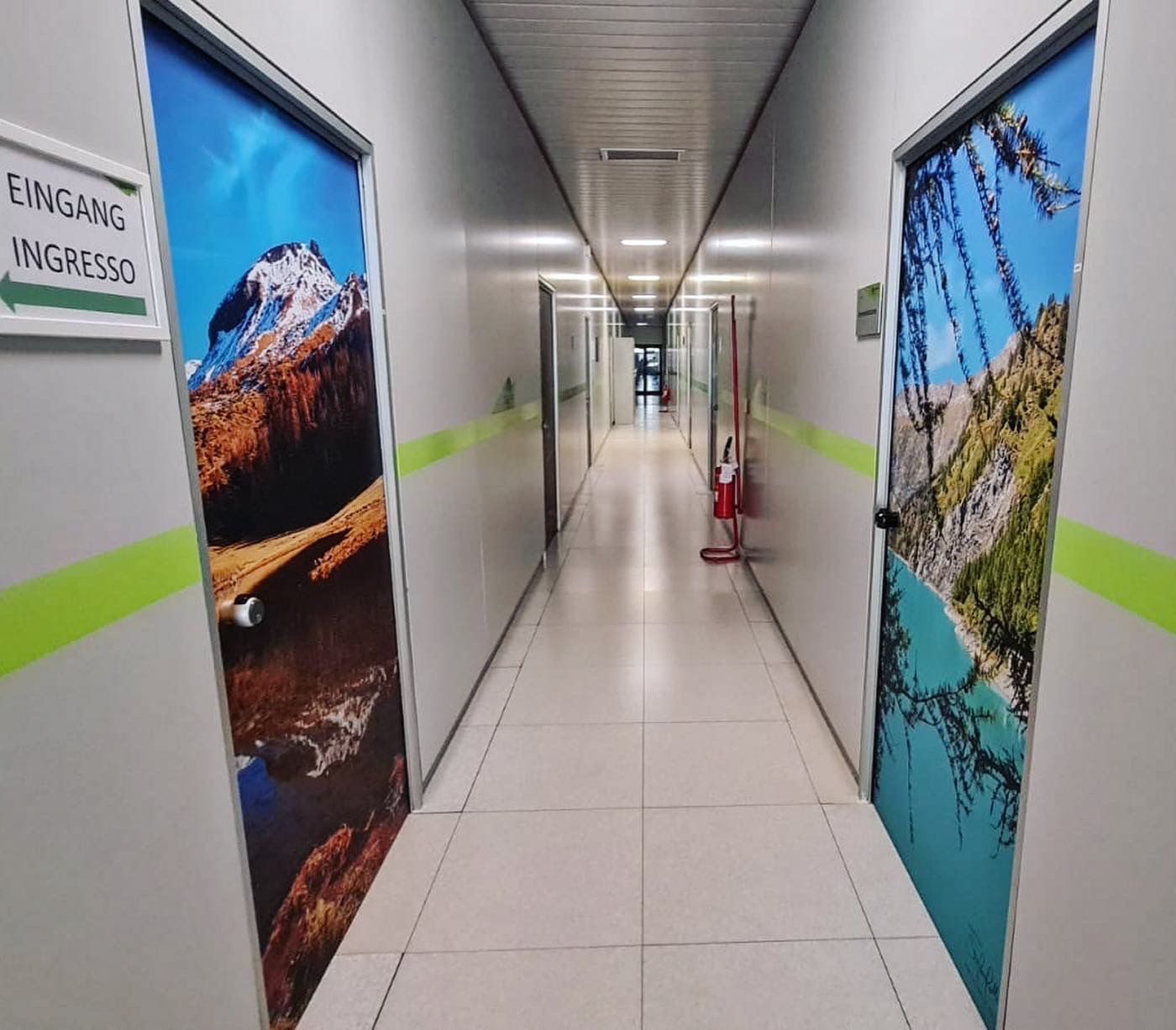 Un corridoio con pareti bianche e pavimento piastrellato, caratterizzato da due porte decorate con grandi stampe a figura intera di montagne e laghi.
