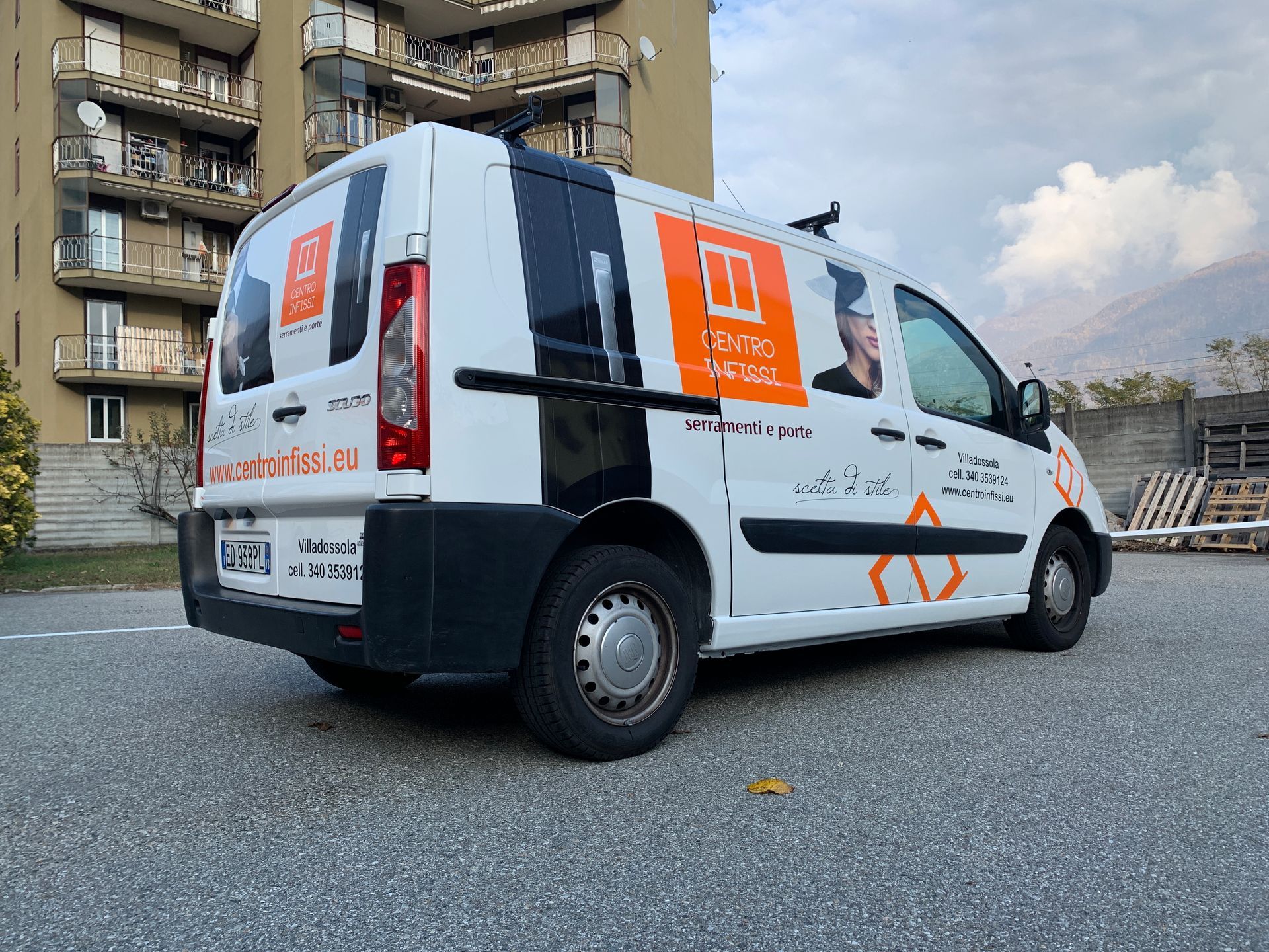 Un furgone commerciale bianco con scritte arancioni e nere, parcheggiato su un piazzale ghiaioso di fronte a un condominio.