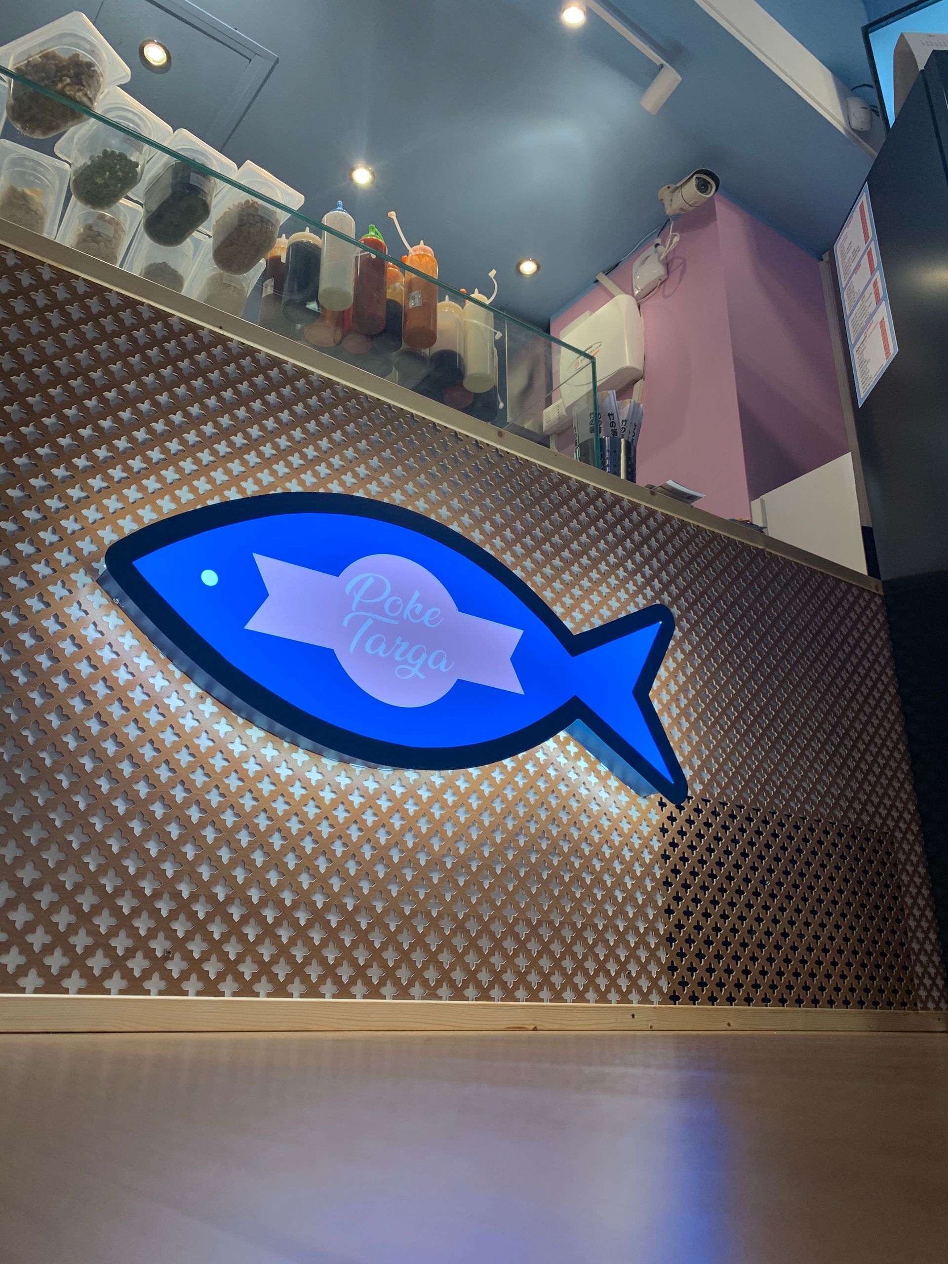 All'interno di un negozio, su una parete marrone con motivo a griglia, è affisso un cartello blu brillante a forma di pesce con testo bianco.