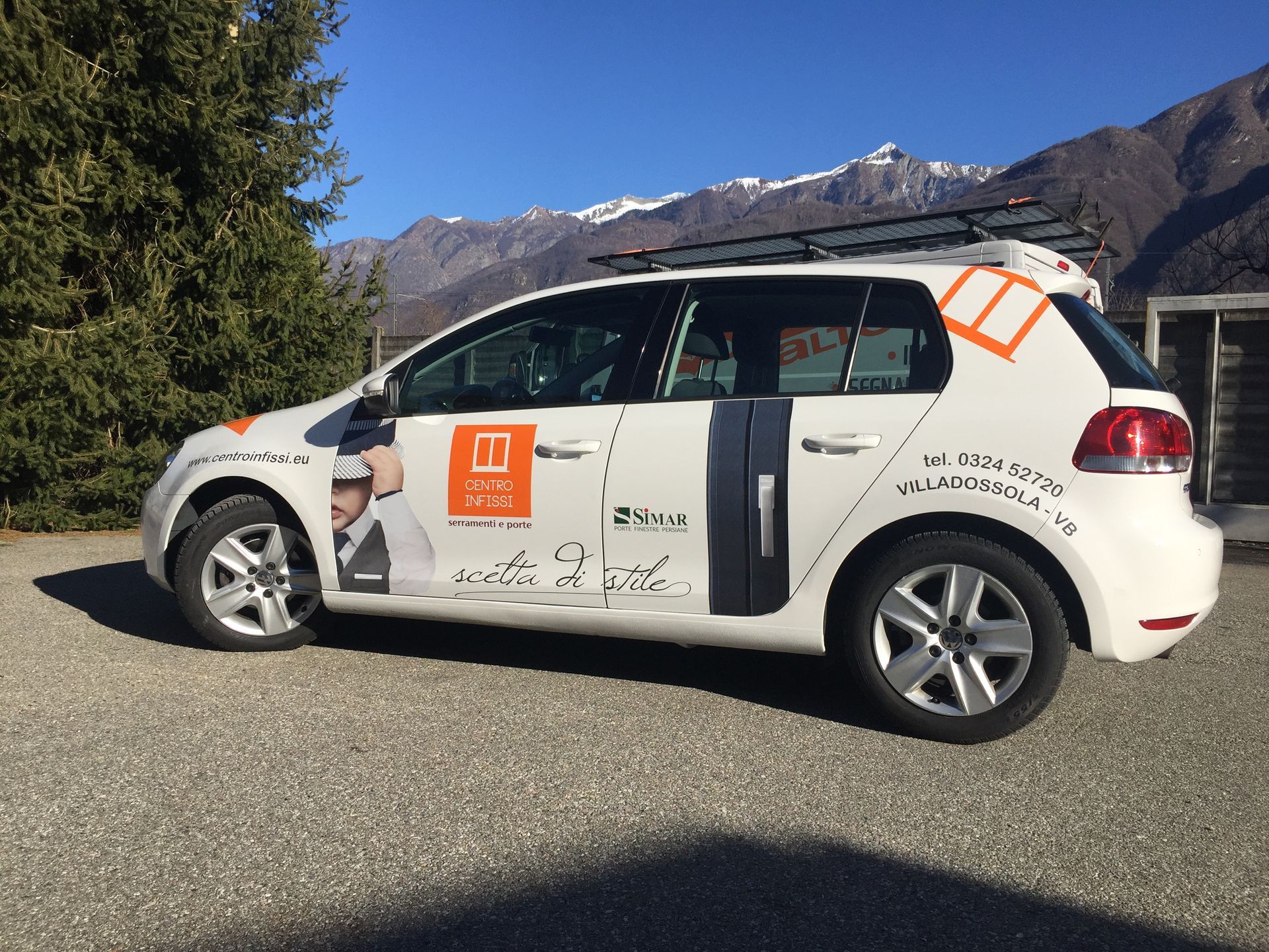 Una Volkswagen hatchback bianca parcheggiata su ghiaia con vista sulle montagne, con il logo promozionale sul lato.