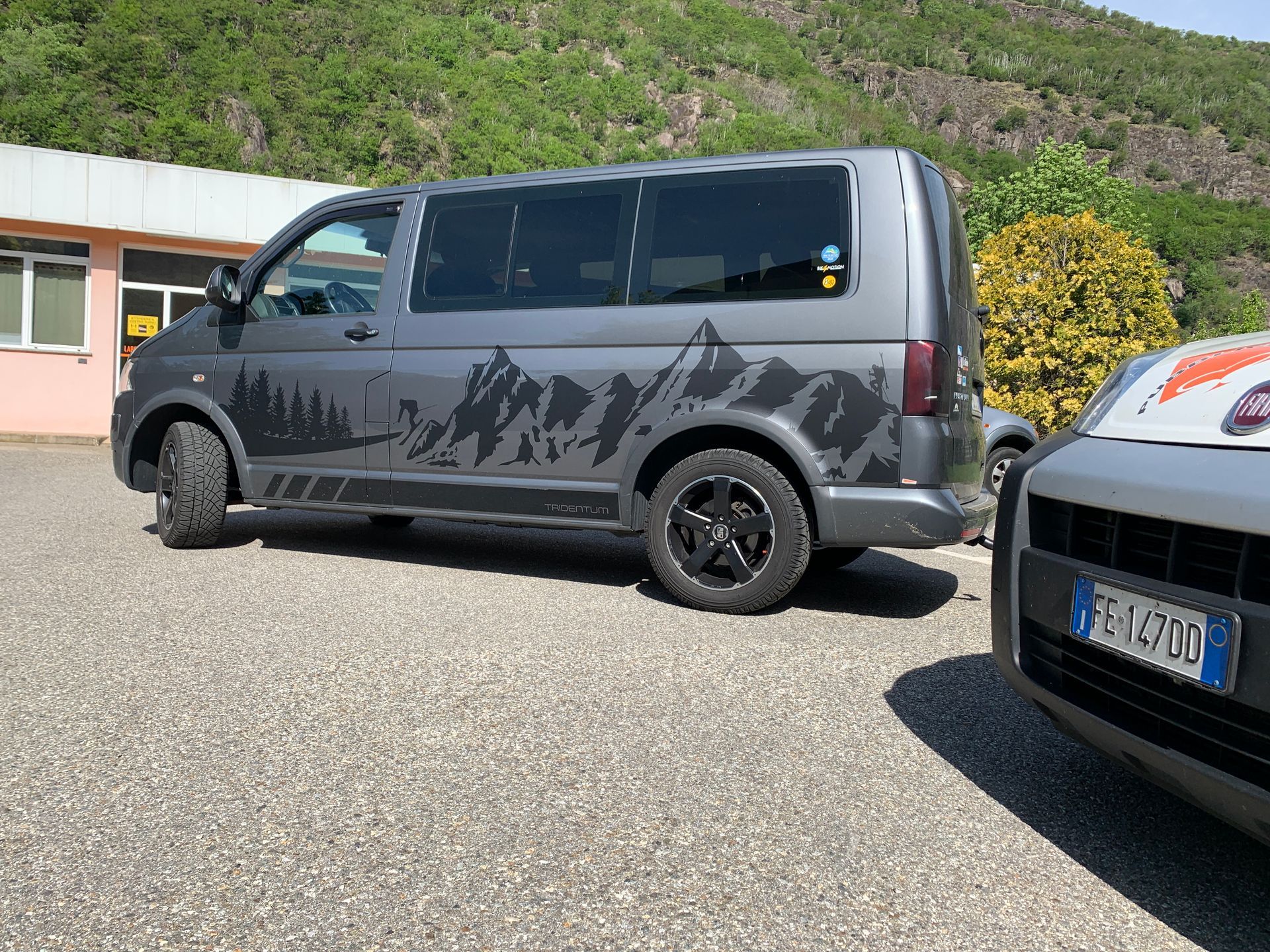 Un furgone Volkswagen Transporter grigio con una decalcomania raffigurante la sagoma di una montagna, parcheggiato su un piazzale ghiaioso vicino a un edificio.