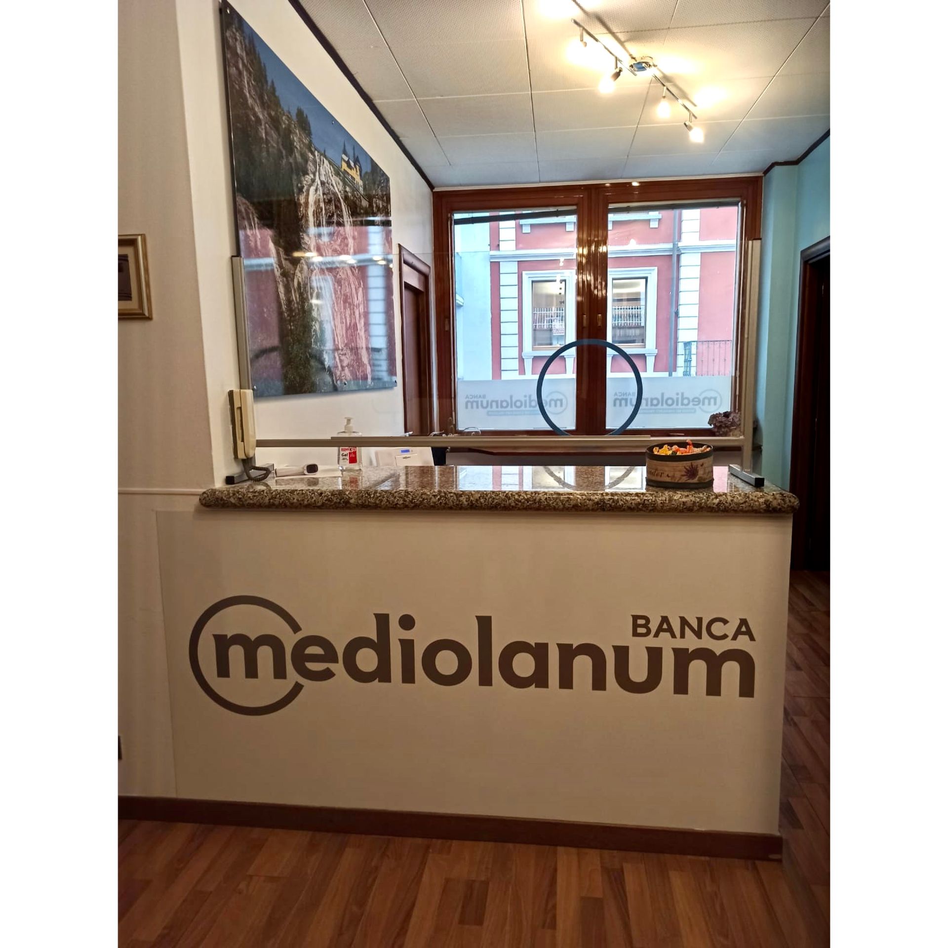 Bancone della reception con il logo della banca Mediolanum, caratterizzato da un piano in pietra e una finestra con vista su un edificio dall'altra parte della strada.