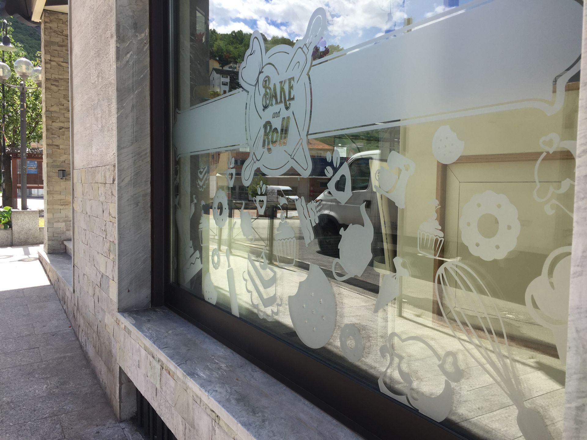 La vetrina di una panetteria presenta adesivi satinati raffiguranti vari prodotti da forno, una frusta e un logo con la scritta 