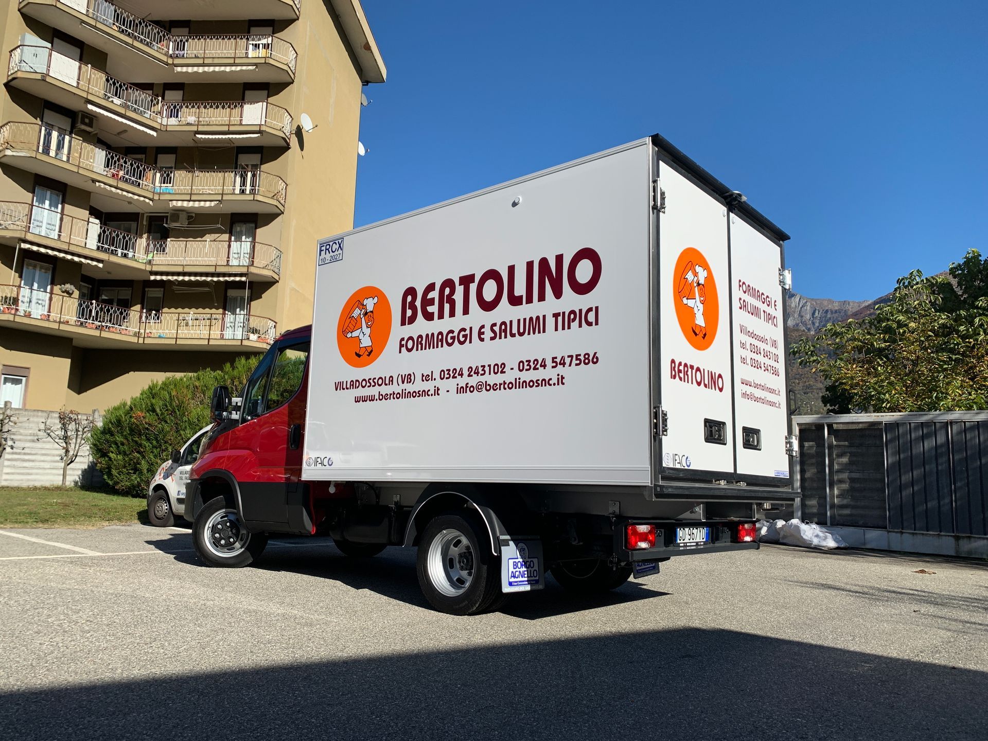 Un furgone per le consegne Bertolino, rosso e bianco, parcheggiato all'aperto su un piazzale ghiaioso accanto a un condominio a più piani.