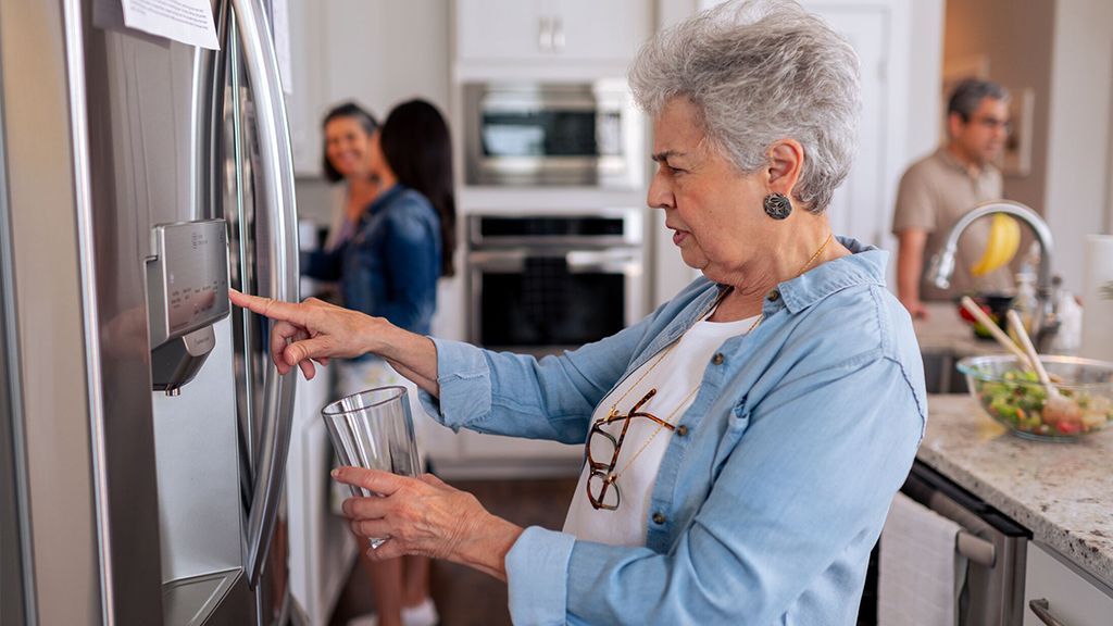 An elderly woman is filling a glass of water from a refrigerator.