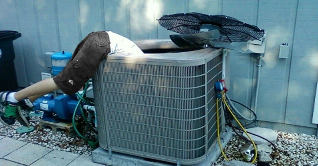 A man is lying on top of an air conditioner trying to repair the inside of the unit. John's Heating, Cooling, and Appliance Repair