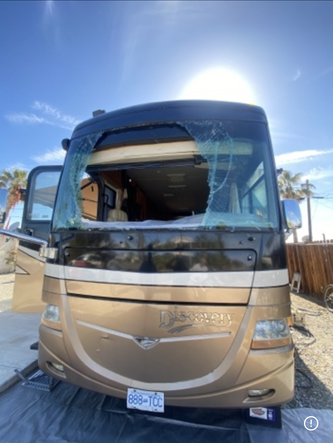 Tan RV with shattered windshield parked outside on a sunny day.