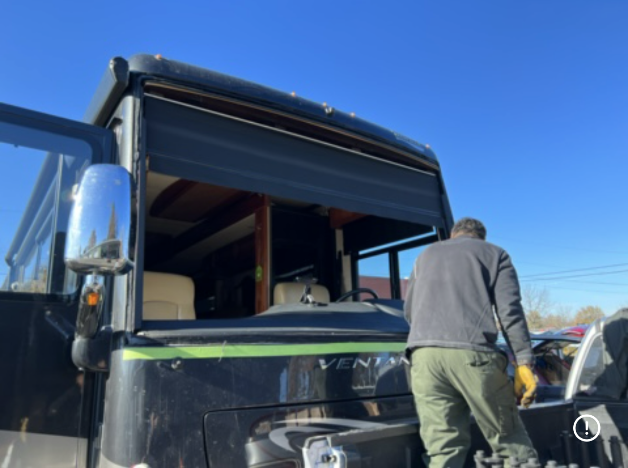 Man working on a black RV windshield with the window removed, outdoors on a sunny day.