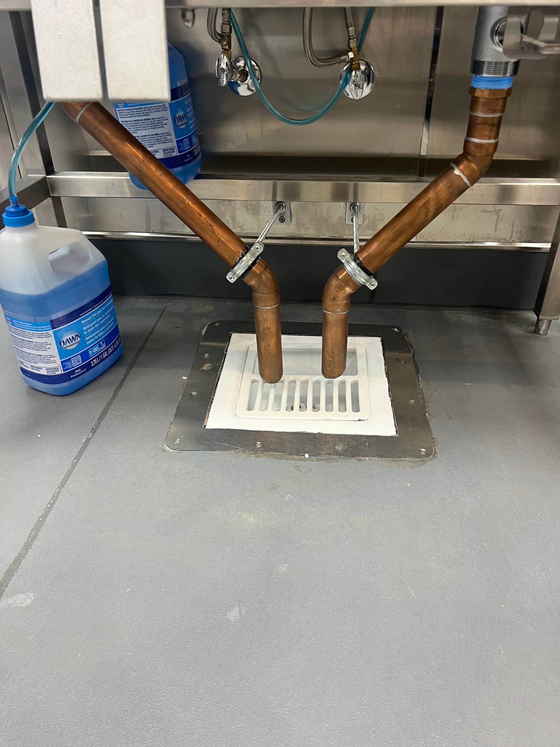 Industrial sink with two copper pipes draining into a floor grate beside a water jug and cleaning cloth.
