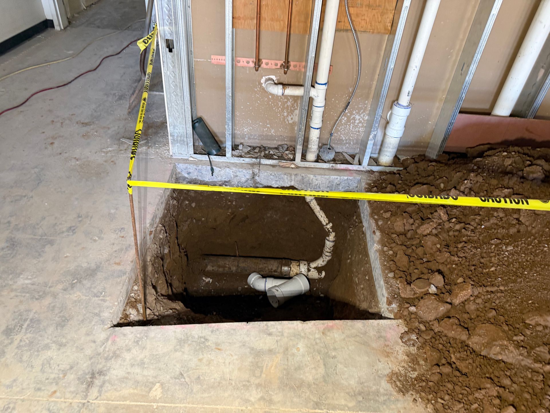 Open floor access hole beside a wall framed with yellow caution tape and exposed plumbing pipes.