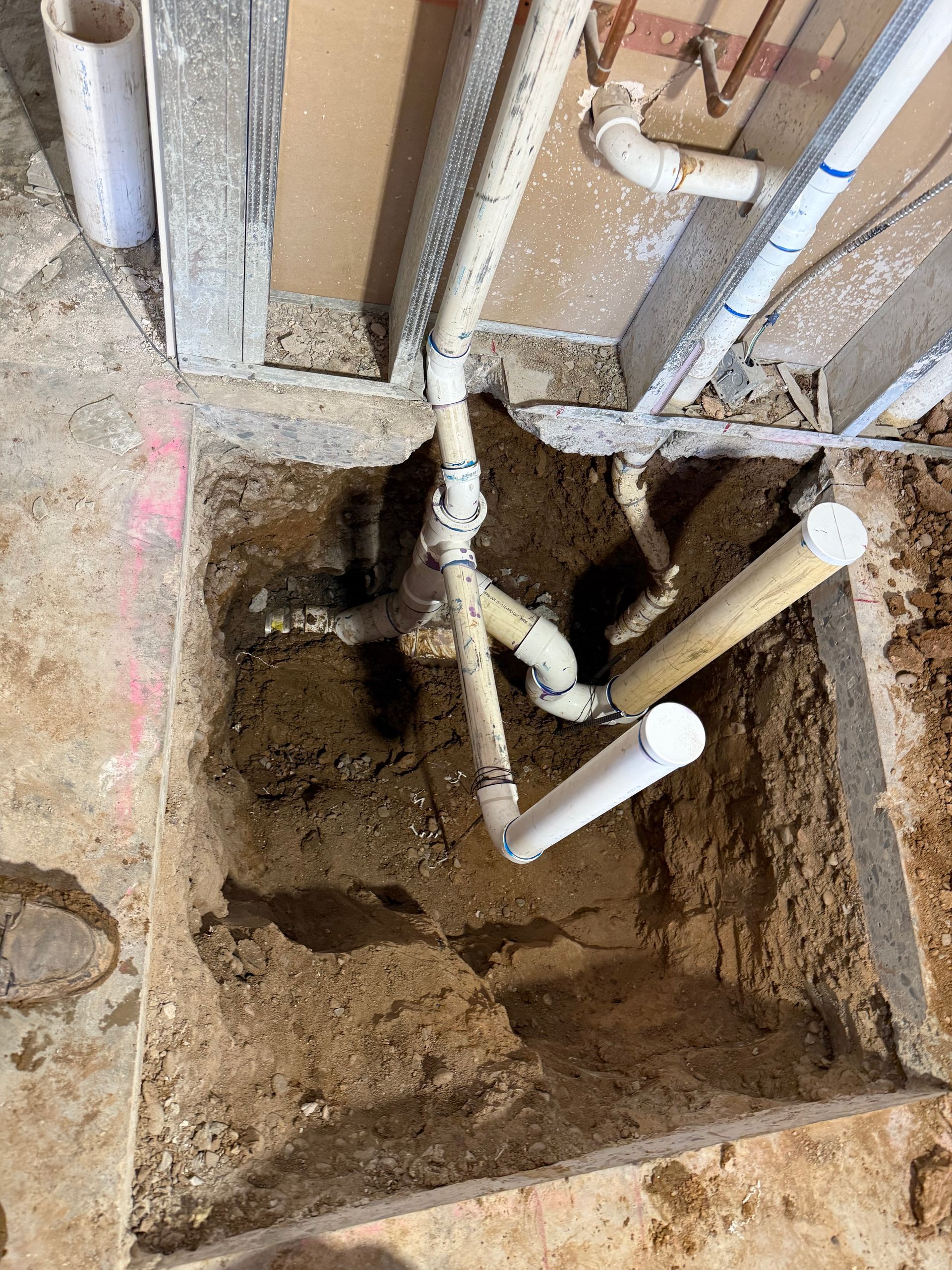 Open floor cavity with exposed plumbing pipes and dirt beneath a wall cutout