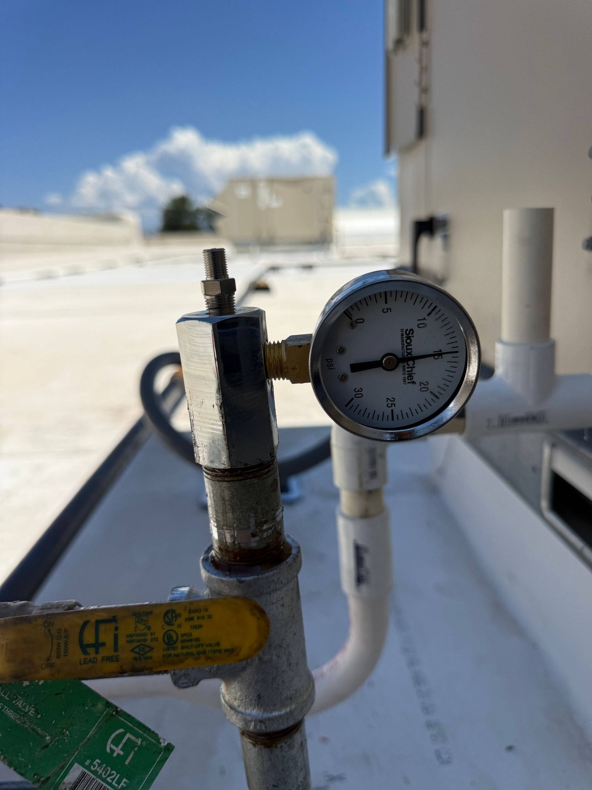 Pressure gauge on a pipe valve outdoors beside a white building, with blue sky in the background.