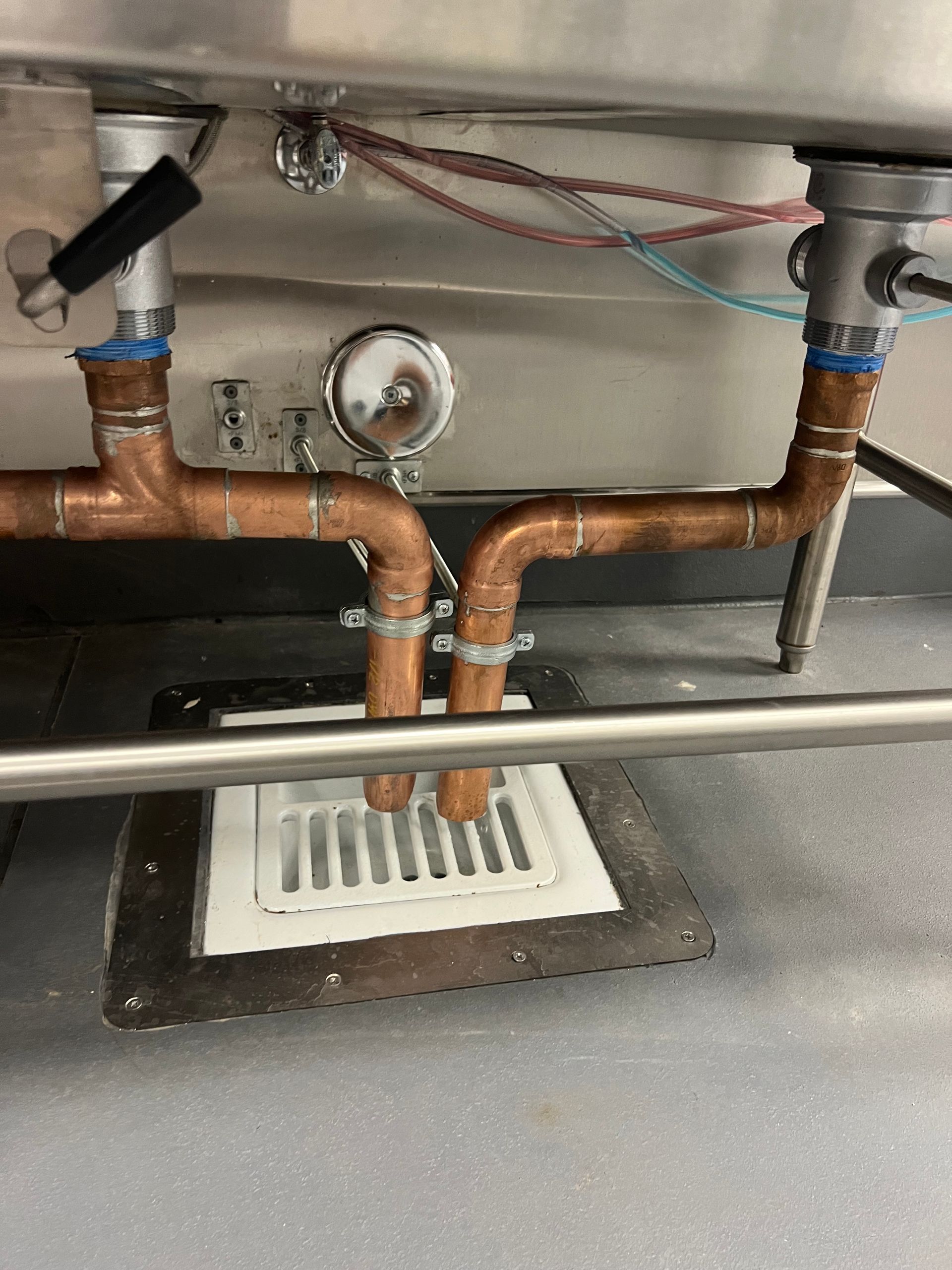 Under-sink copper plumbing with a floor drain and white grate, showing pipes, valves, and a pressure gauge