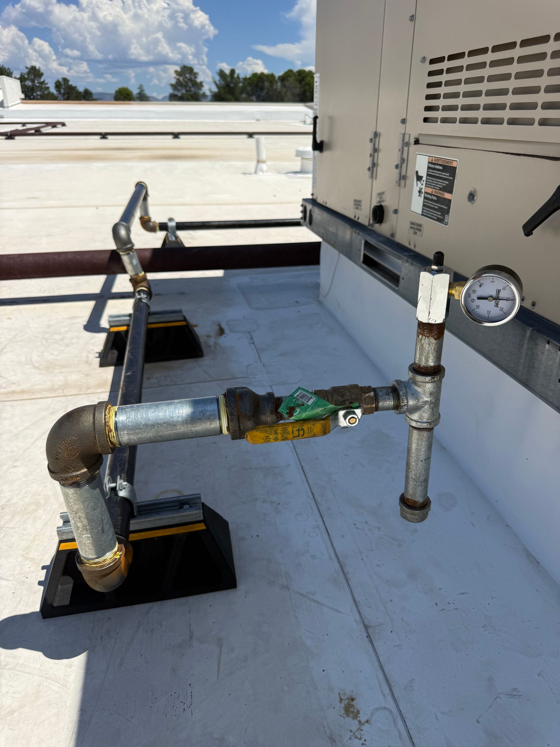 Rooftop industrial pipes and valves with a pressure gauge on a white utility unit.