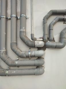 Gray metal pipes with elbow joints running along a wall, secured by clamps and brackets.