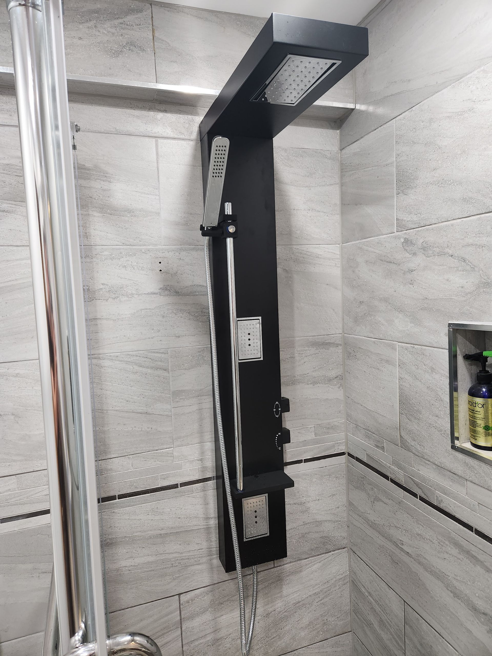 Black shower panel with overhead showerhead and body jets against gray tiled wall.