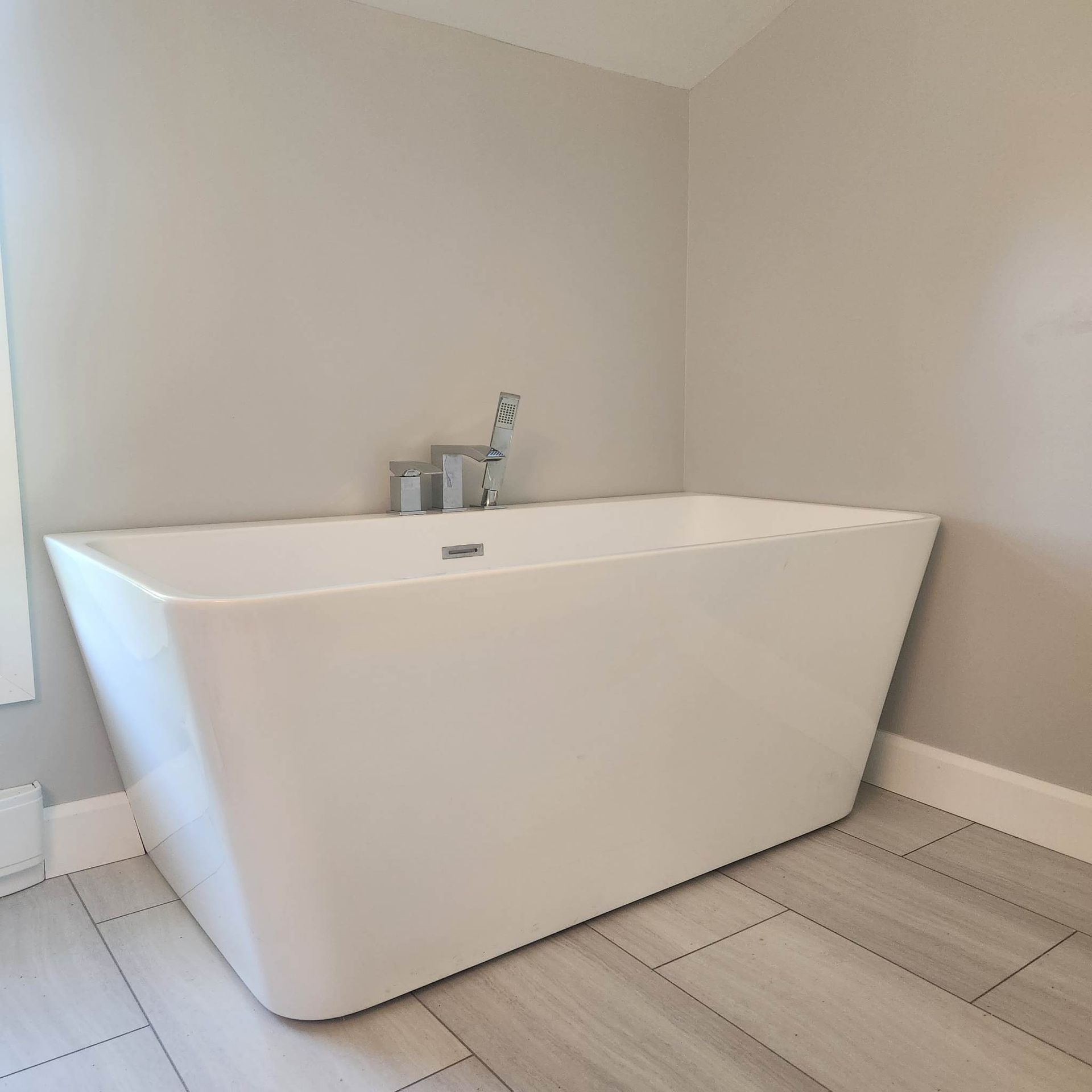 White rectangular bathtub against gray walls and light-colored tiled floor. Faucet and handheld showerhead are visible.