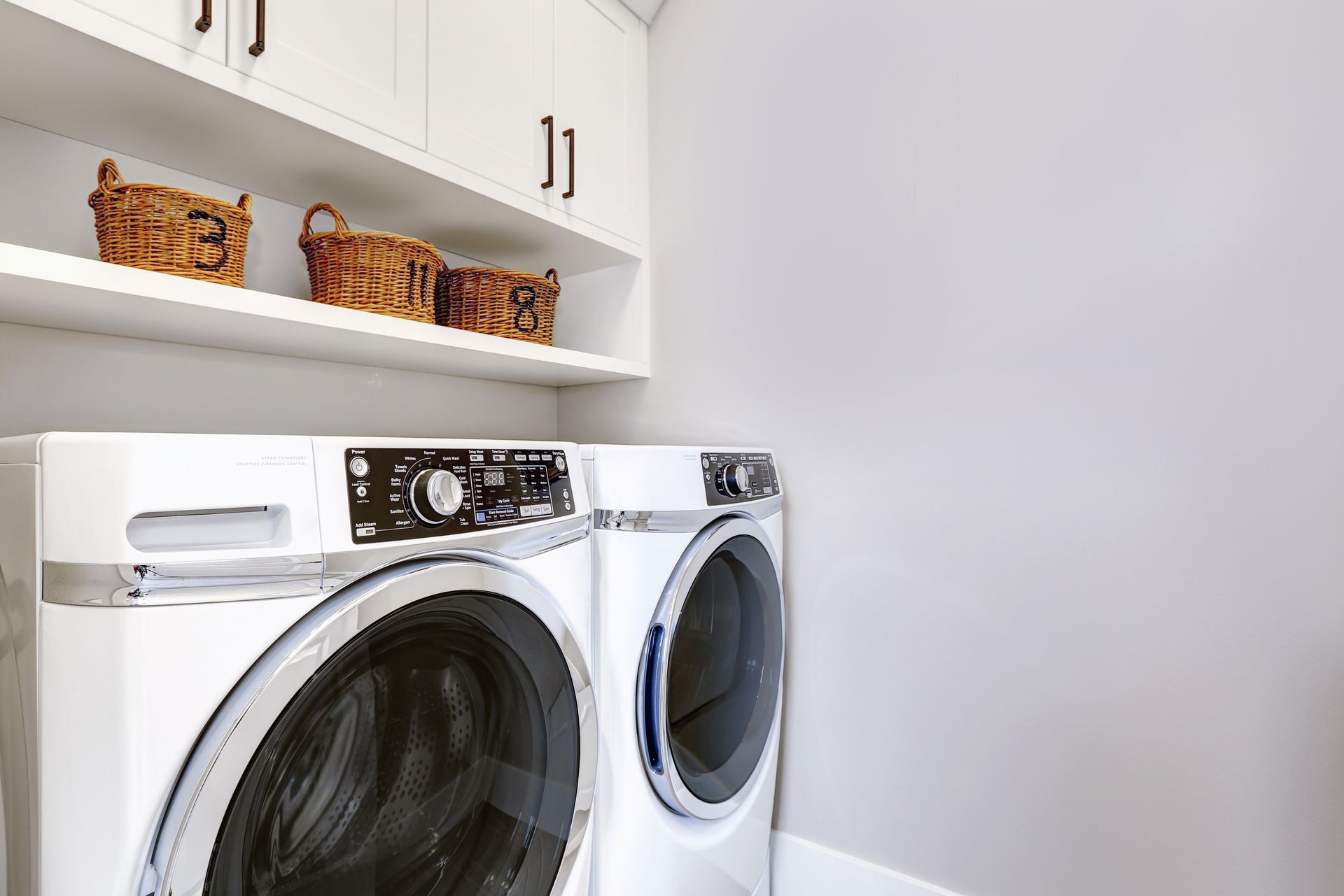 A laundry room features two white front-load appliances beneath a white shelf holding three wicker baskets and cabinets.