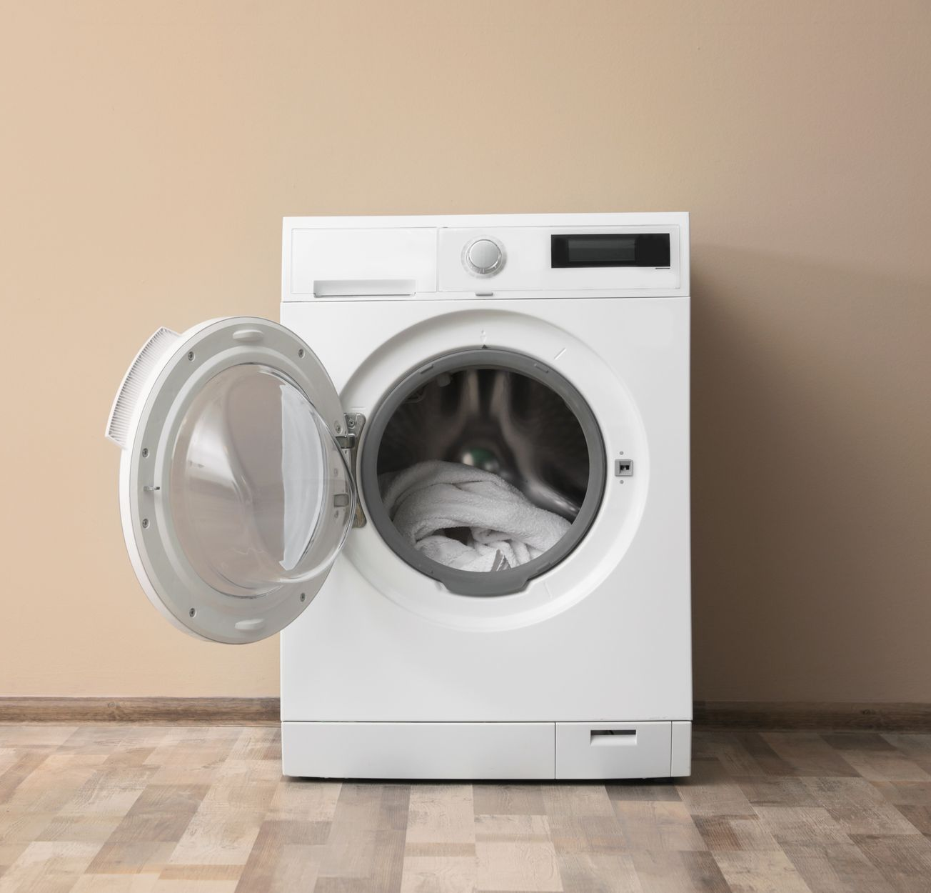 A white front-loading washing machine with its door open, standing on a wooden floor against a beige wall.
