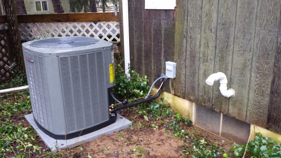 Outdoor air conditioner unit beside a wooden house wall, with a vent pipe and electrical line nearby