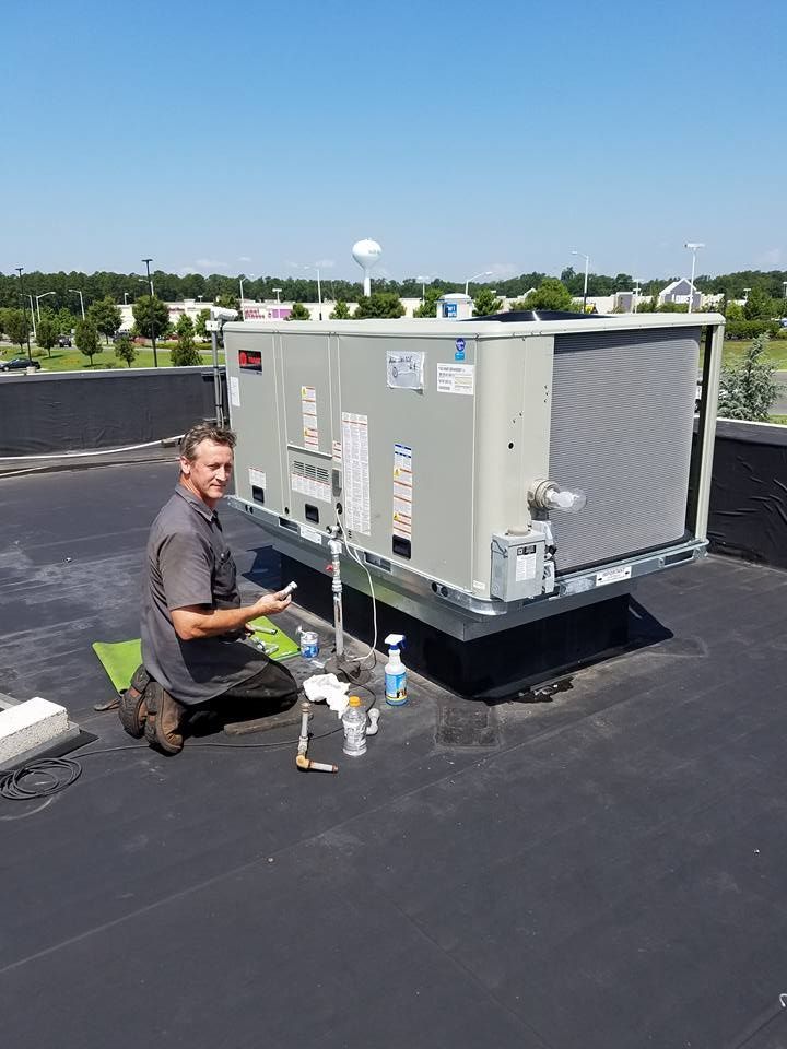 Climate Control Services technician kneeling beside rooftop HVAC unit, inspecting equipment on a sunny day