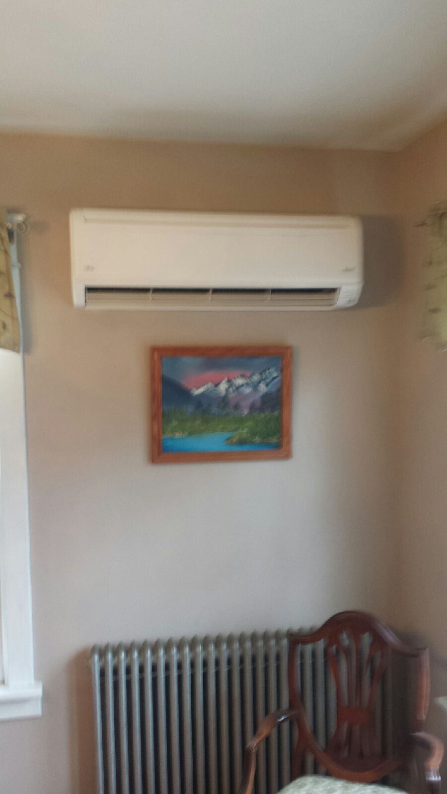 Wall-mounted air conditioner above a framed landscape painting, with a radiator and wooden chair below