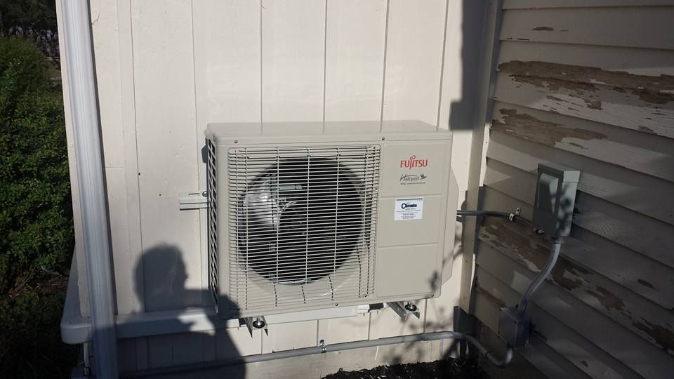 Outdoor Fujitsu HVAC condenser unit beside a white house, with hoses and a shadow in late-afternoon sun