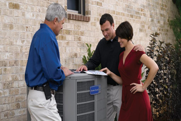 Three people inspecting an outdoor air conditioner by a brick house, with one pointing at paperwork
