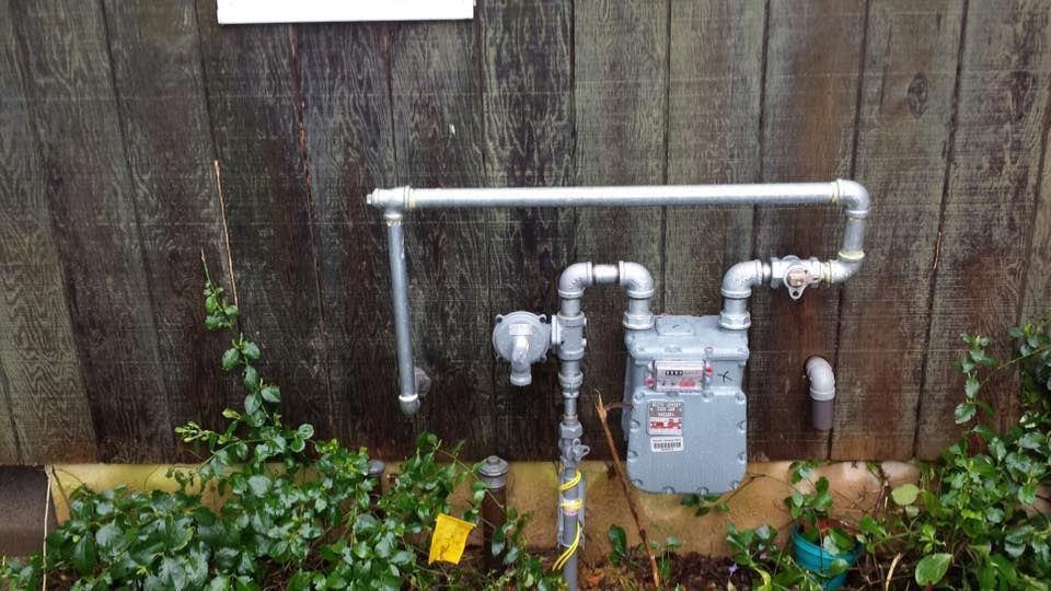 Gas meter and white pipes mounted on a wooden fence above garden plants