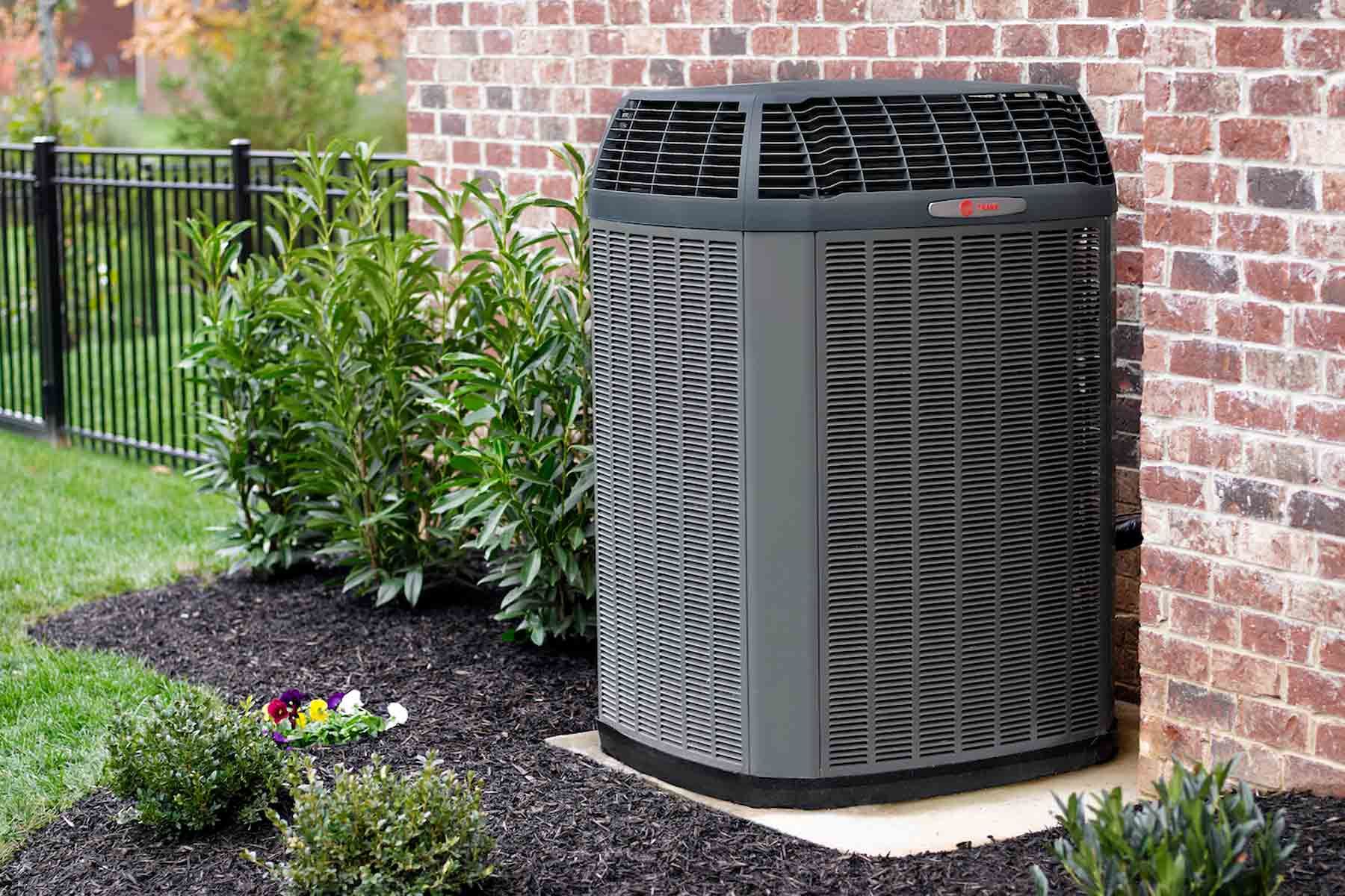 Two outdoor HVAC units beside plants, with black grilles and gray tops on a concrete pad