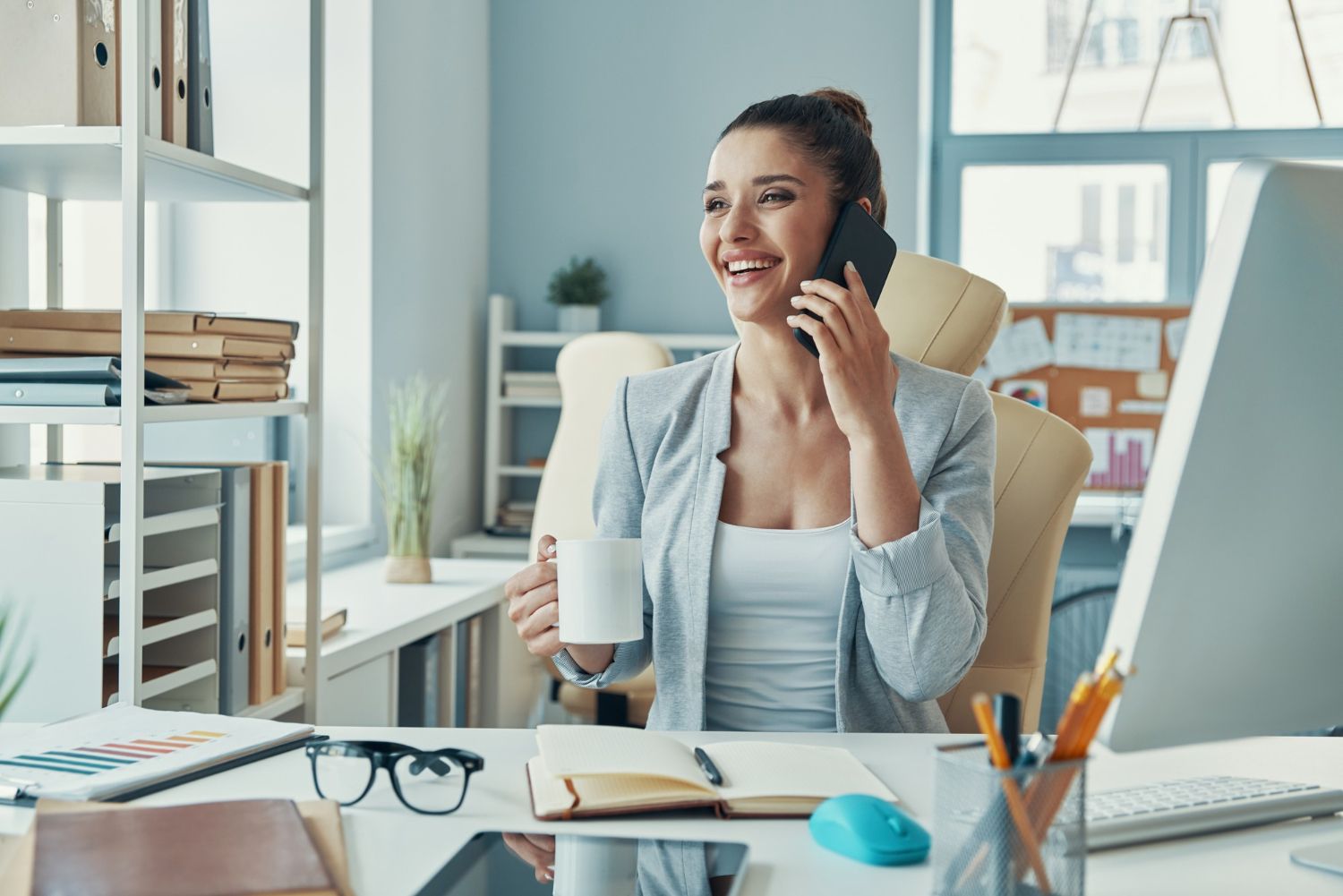 Woman smiling on a phone call at a desk in a bright office, holding a mug and seated by a laptop.