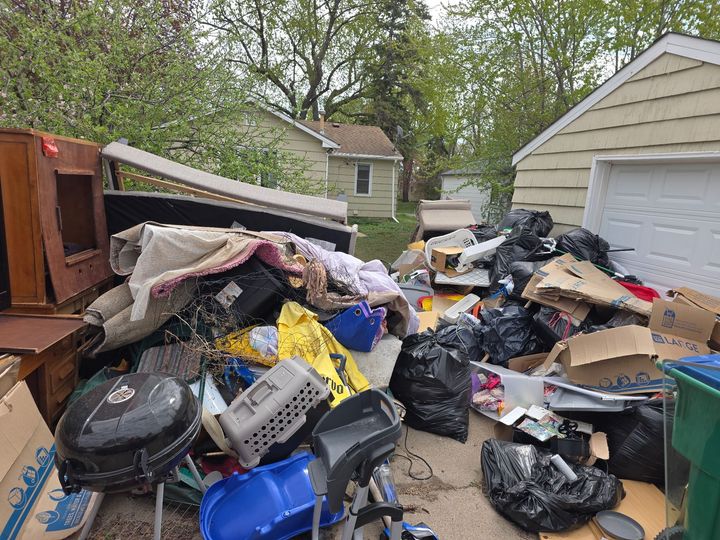 Cluttered driveway piled with trash bags, broken furniture, and debris beside a garage