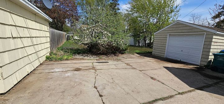 Concrete driveway beside a beige house and detached garage, with trees and a clear blue sky.