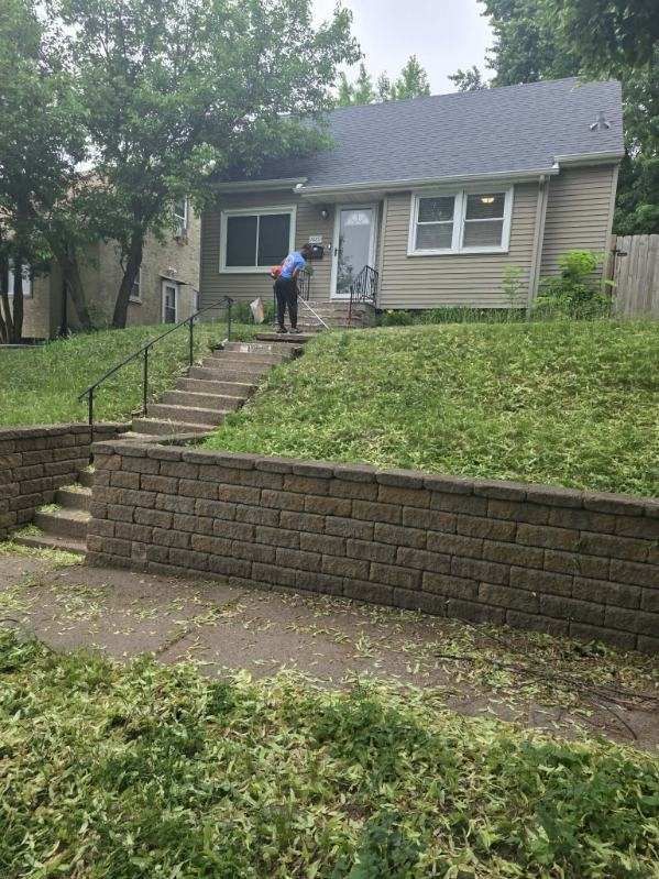 Person walking up steps to a small house on a grassy slope with retaining walls and trees.