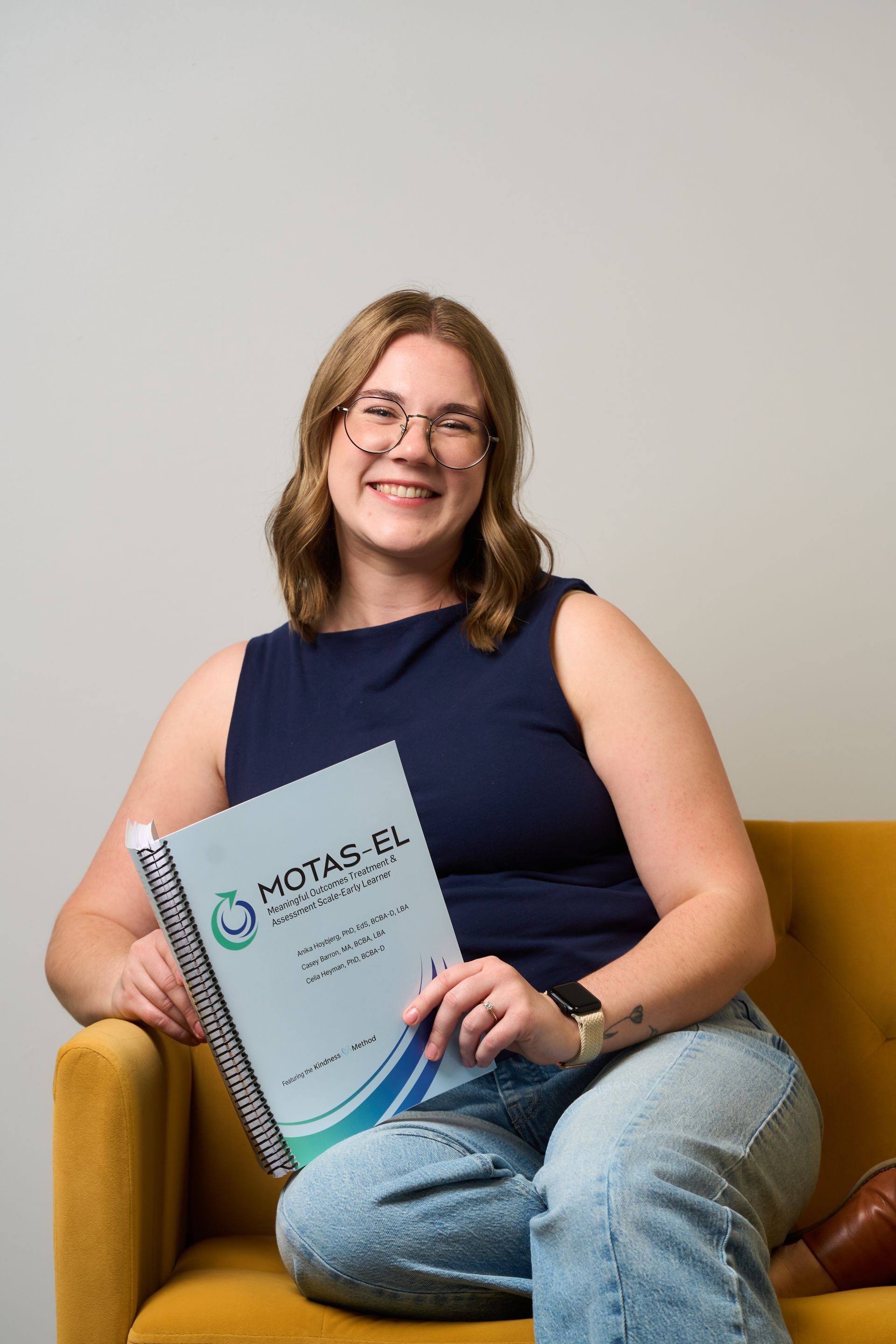 Woman seated on a mustard chair holding a certificate, smiling against a plain wall.
