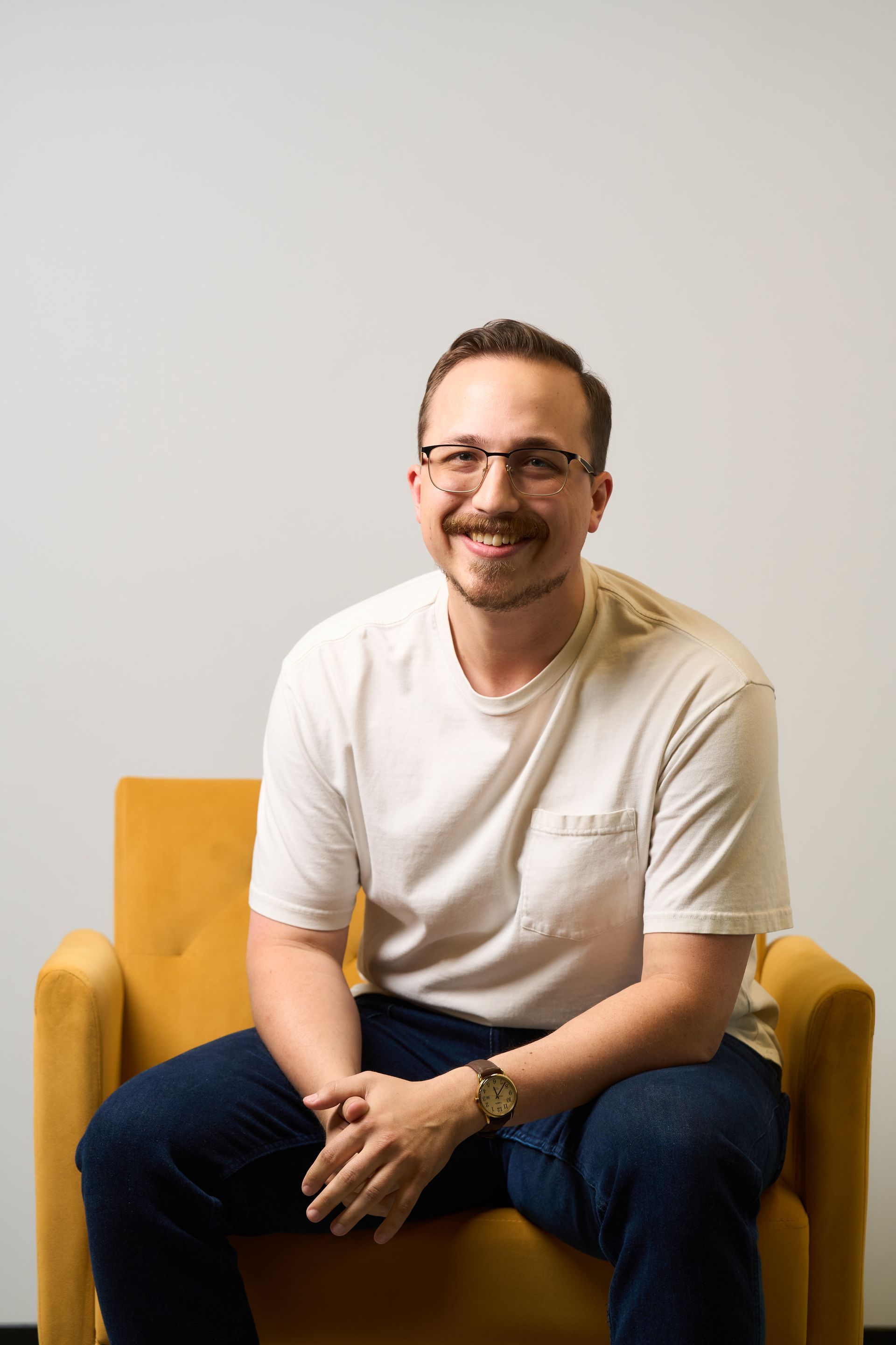 Man seated in a mustard yellow chair against a light wall, smiling and wearing glasses, a white shirt, and dark pants.