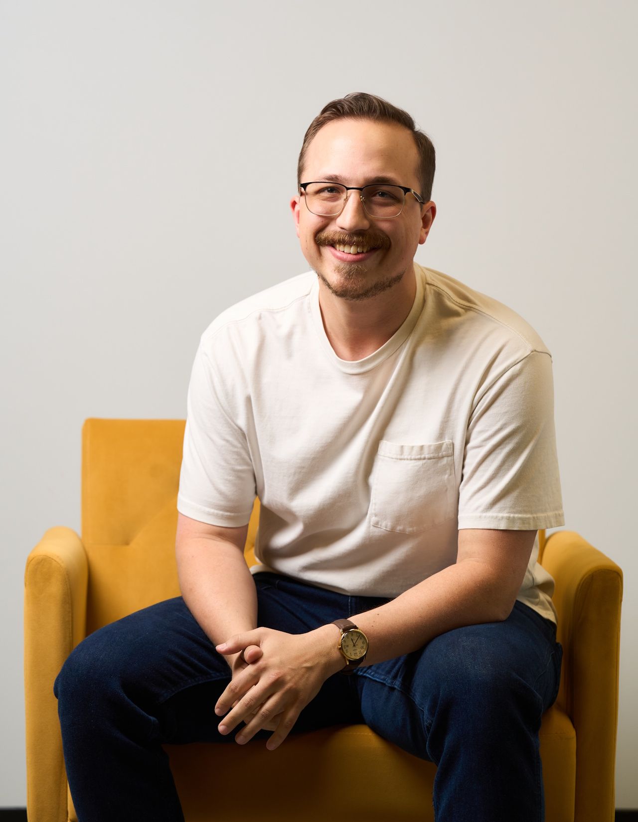 Man seated in a yellow armchair, smiling, wearing glasses and a light shirt against a plain background.