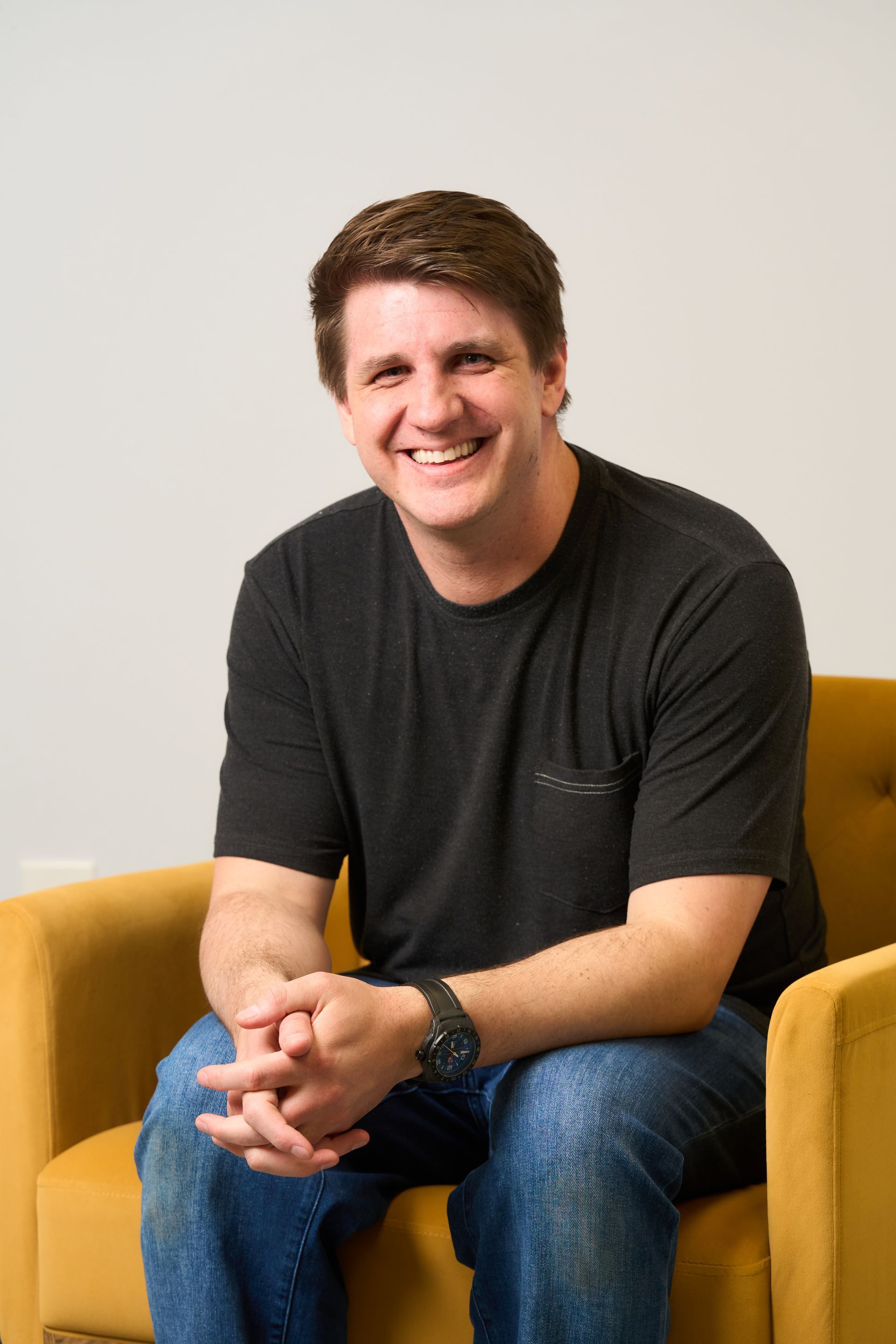Smiling person seated in a mustard-yellow chair, holding a pink tablet against a light background