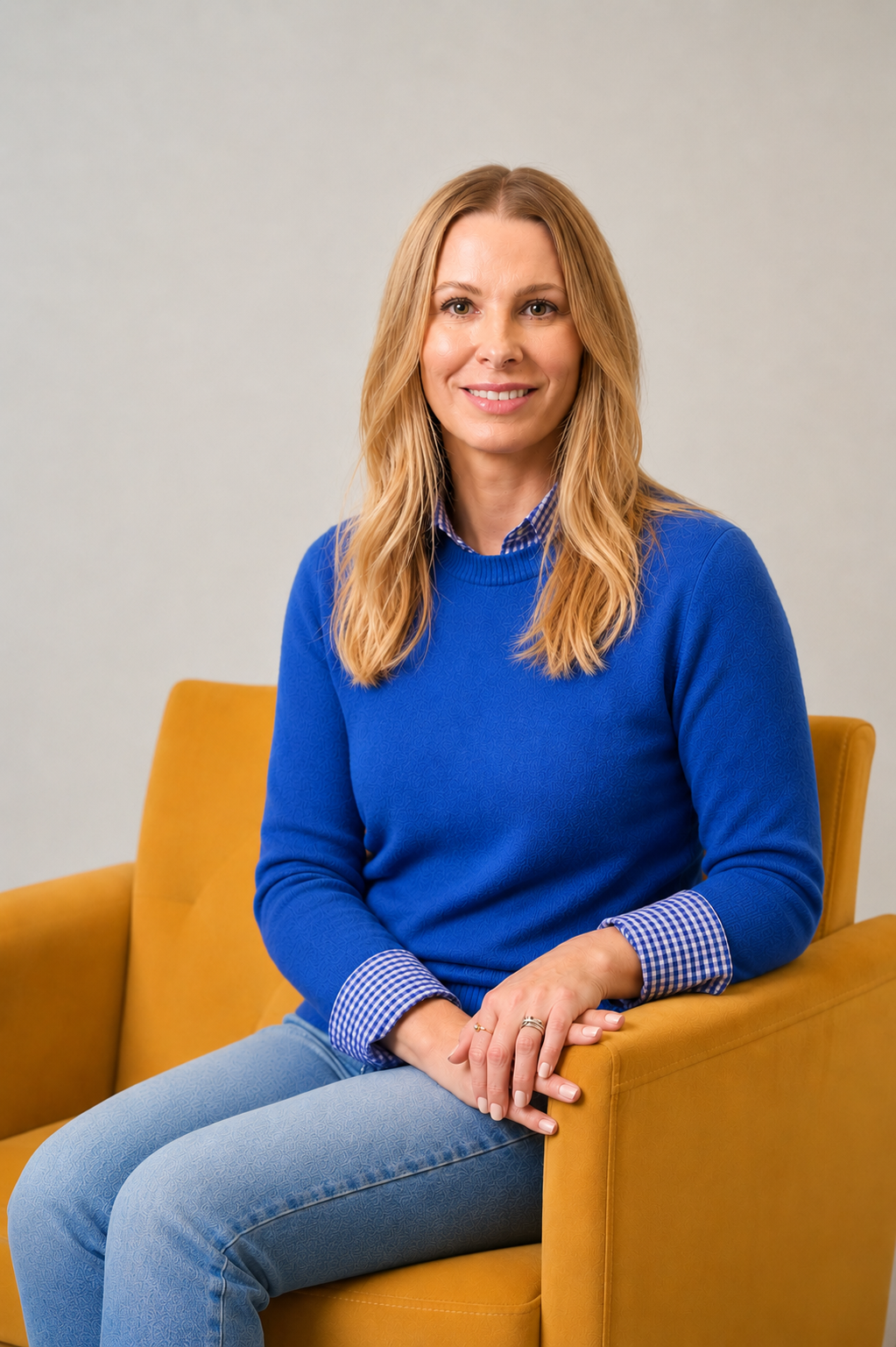 Woman in a blue sweater sitting in a mustard-yellow chair, smiling against a plain background.