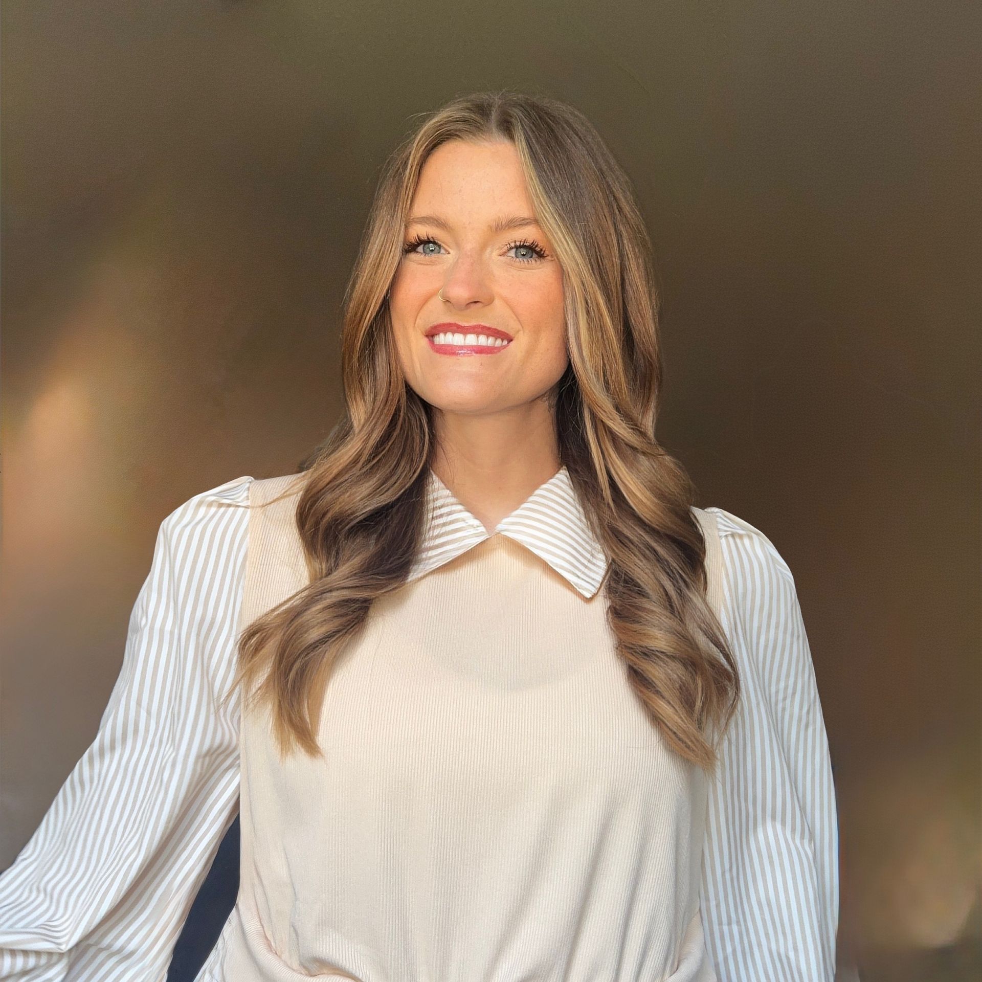 Smiling woman in a white blouse with a wide collar, standing against a warm blurred background