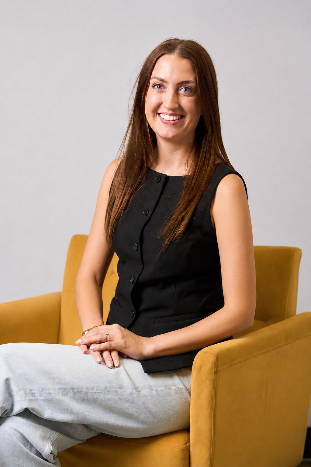 Woman seated in a mustard-yellow chair, smiling in a black sleeveless top and light pants.