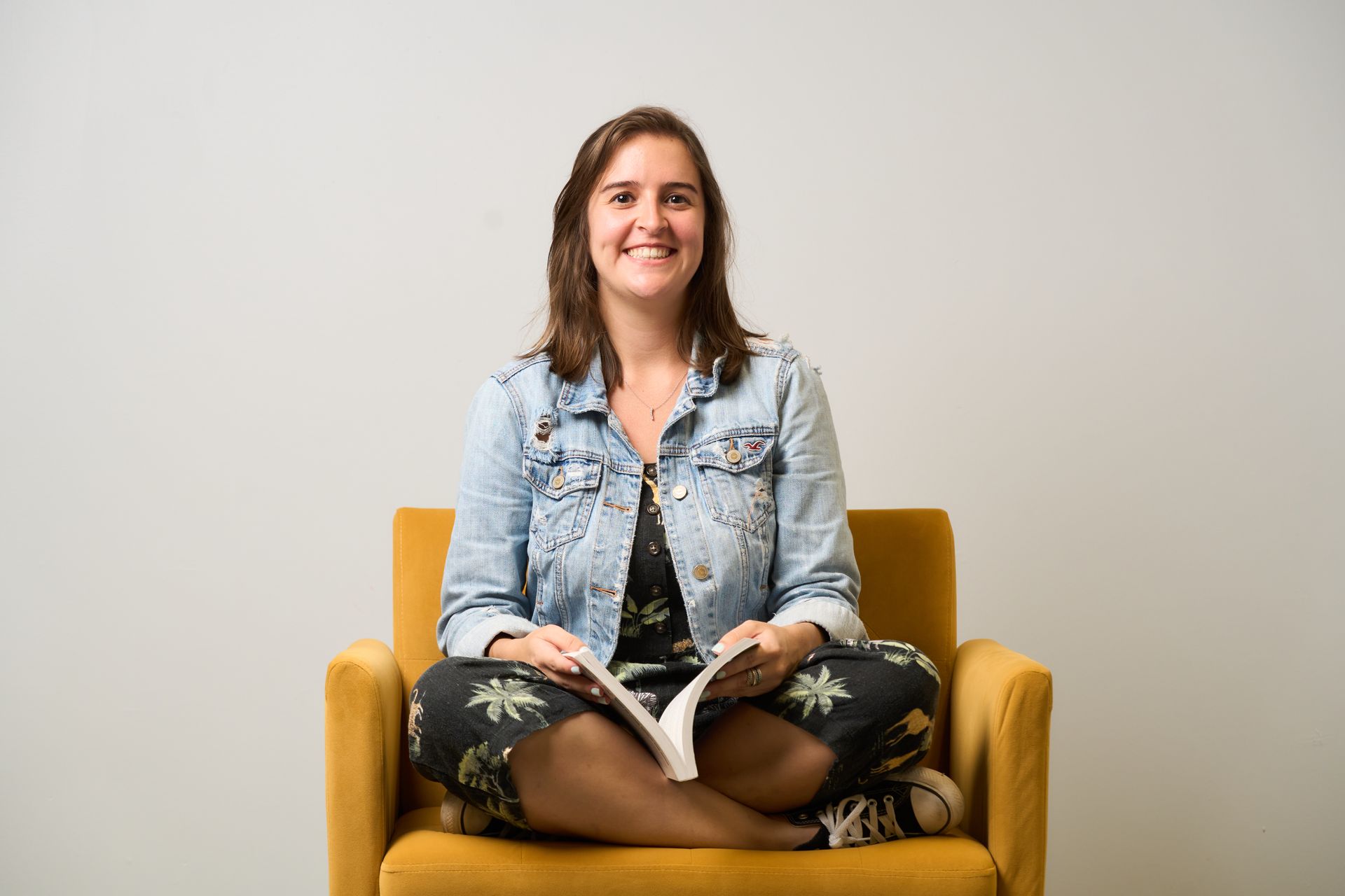 Smiling person seated in a mustard-yellow chair, holding a pink tablet against a light background