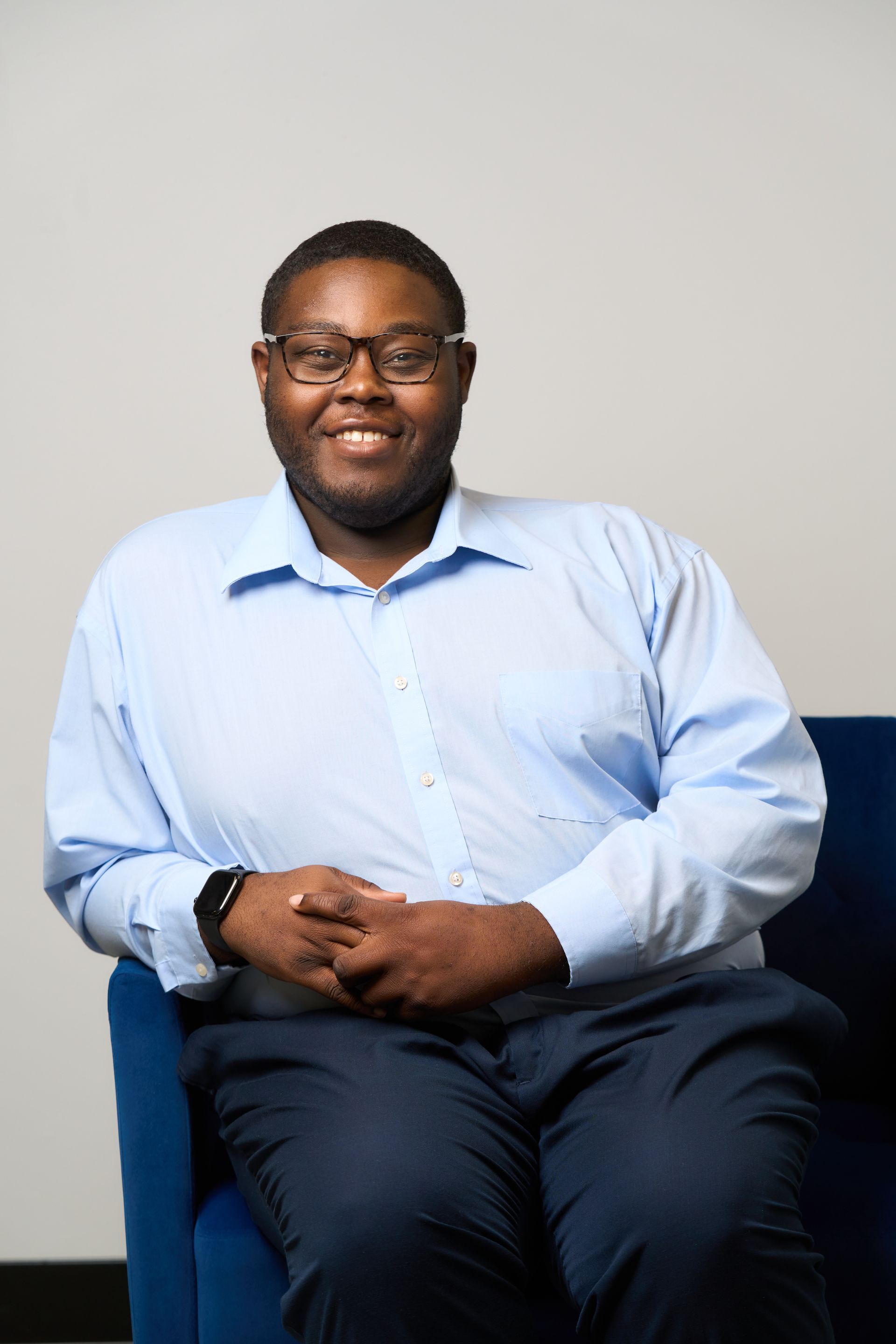 Man seated in a mustard yellow chair against a light wall, smiling and wearing glasses, a white shirt, and dark pants.