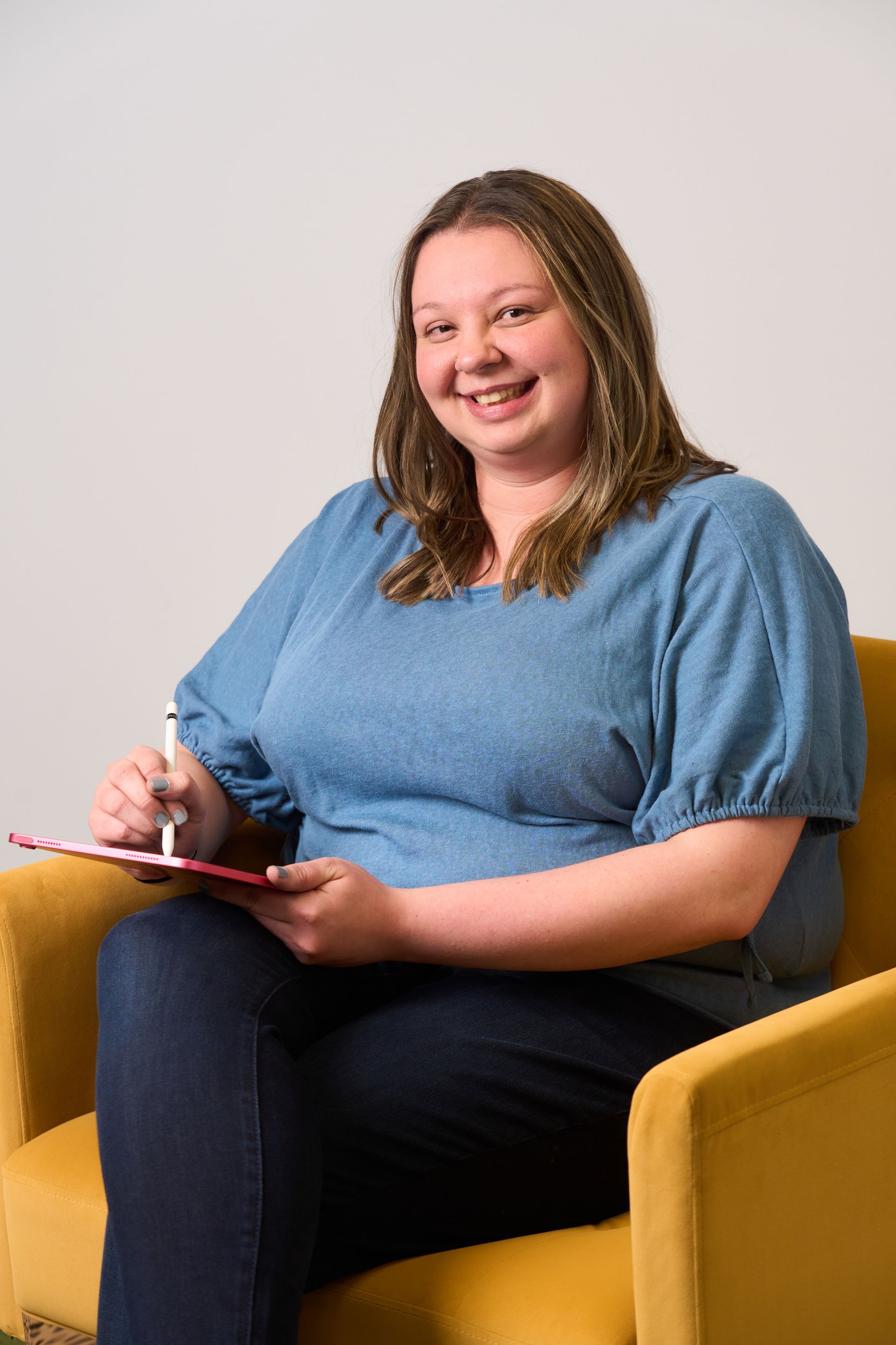 Smiling person seated in a mustard-yellow chair, holding a pink tablet against a light background