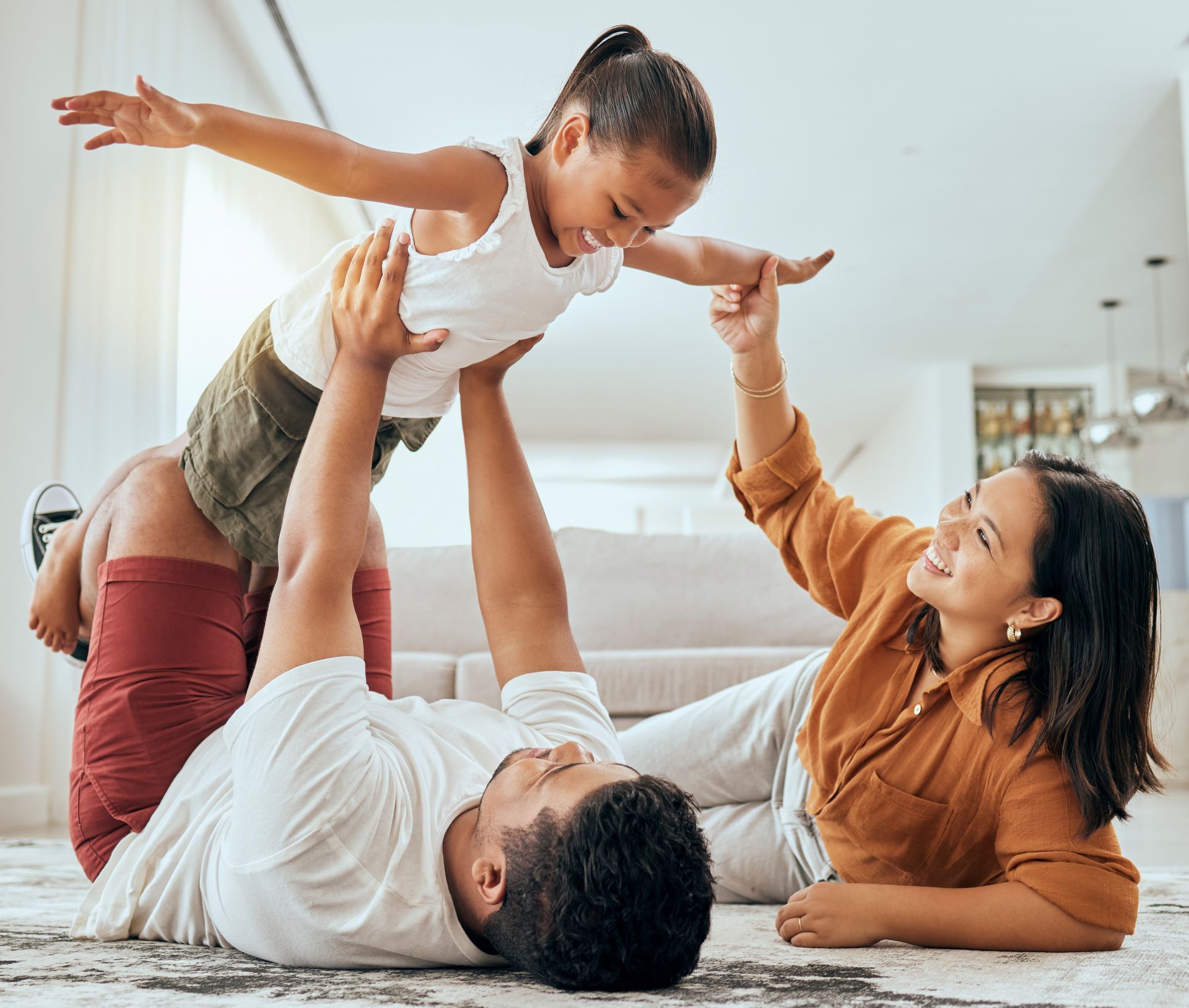 Two adults lifting a smiling child indoors, with a soft, bright background.